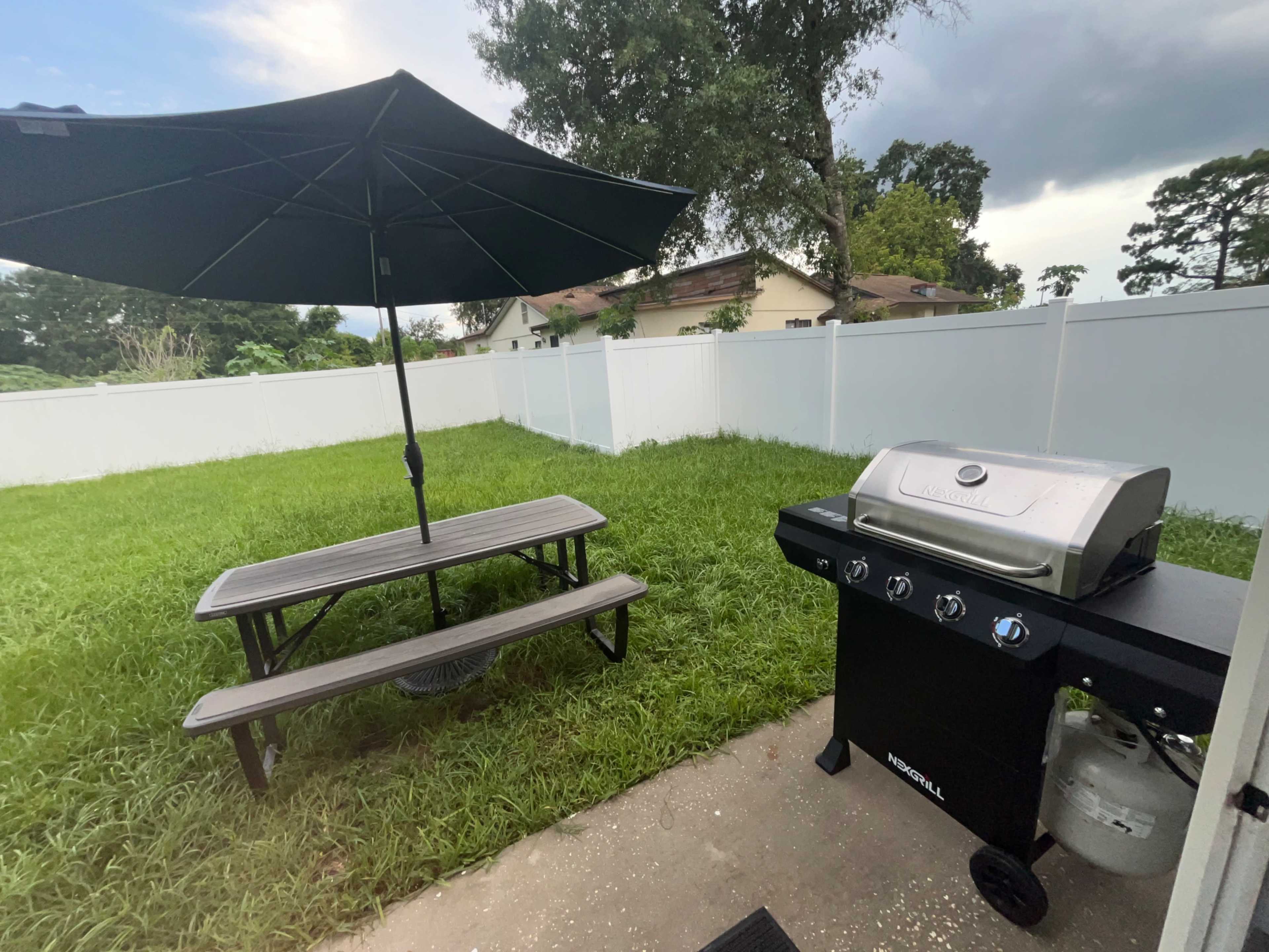 The image shows a barbecue grill beside a picnic table with an umbrella in a yard surrounded by a white fence and tall grass.