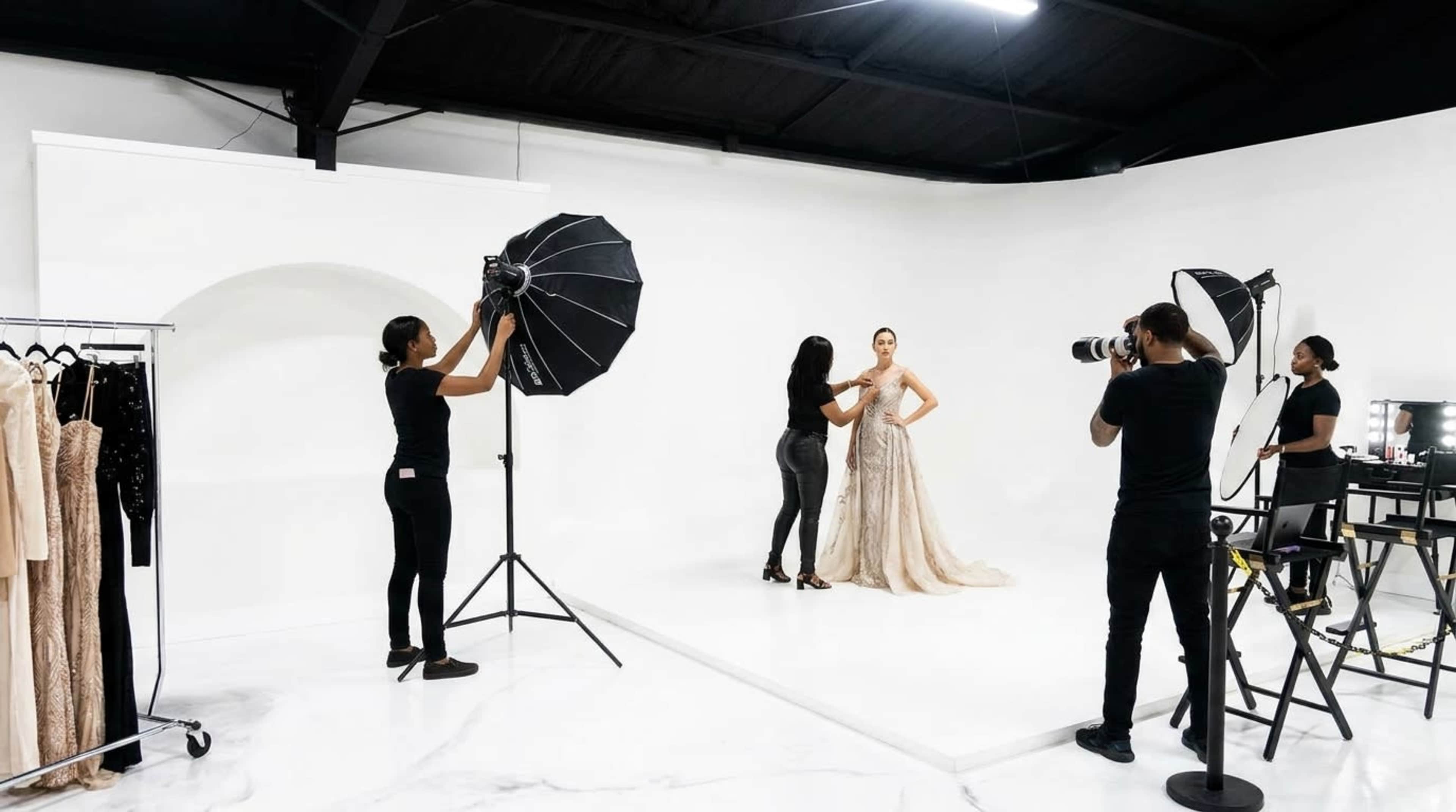 A fashion photoshoot takes place in a well-lit studio where a model is being styled with a flowing gown while a photographer captures the scene.
