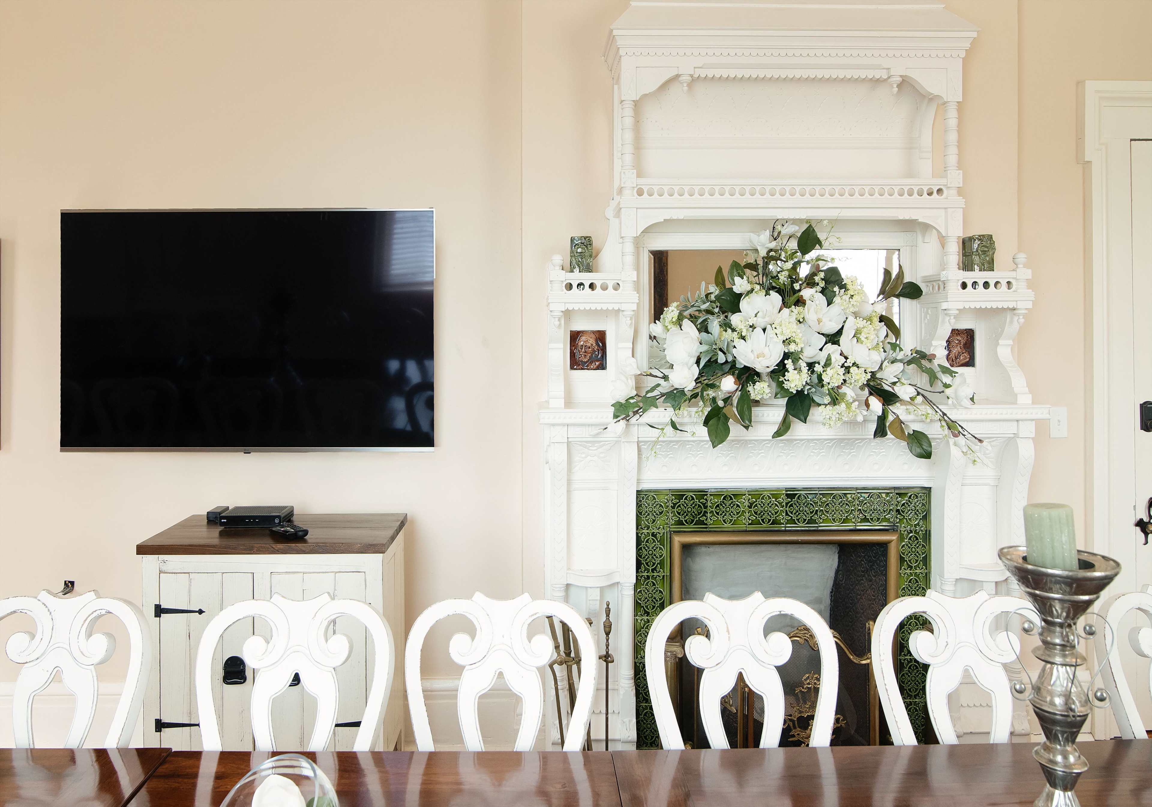 A dining room features a long wooden table with white chairs, a flat-screen TV mounted on the wall, and a decorative fireplace adorned with greenery and flowers.