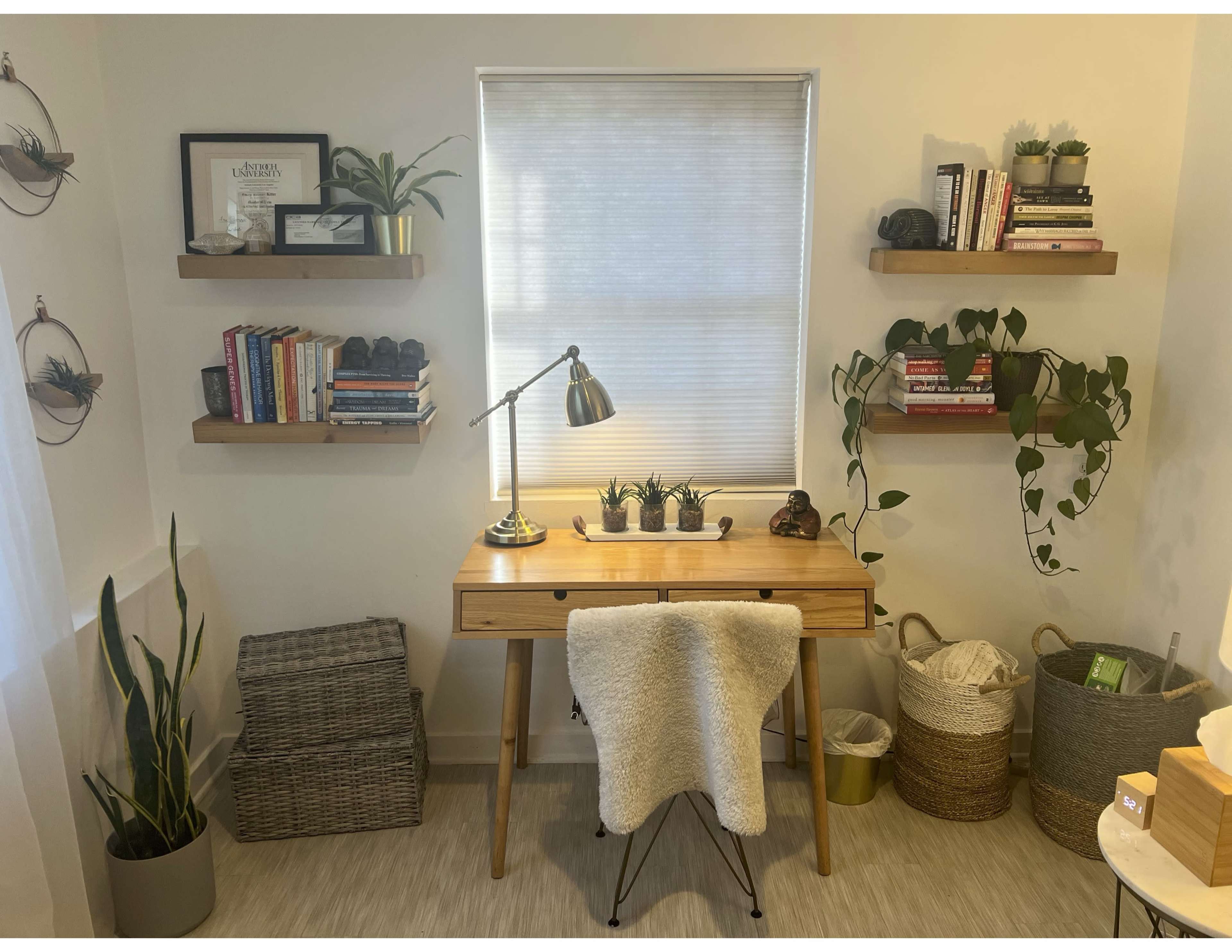 The image shows a small home office with a wooden desk, a lamp, bookshelves, and plants arranged on the walls and surfaces.