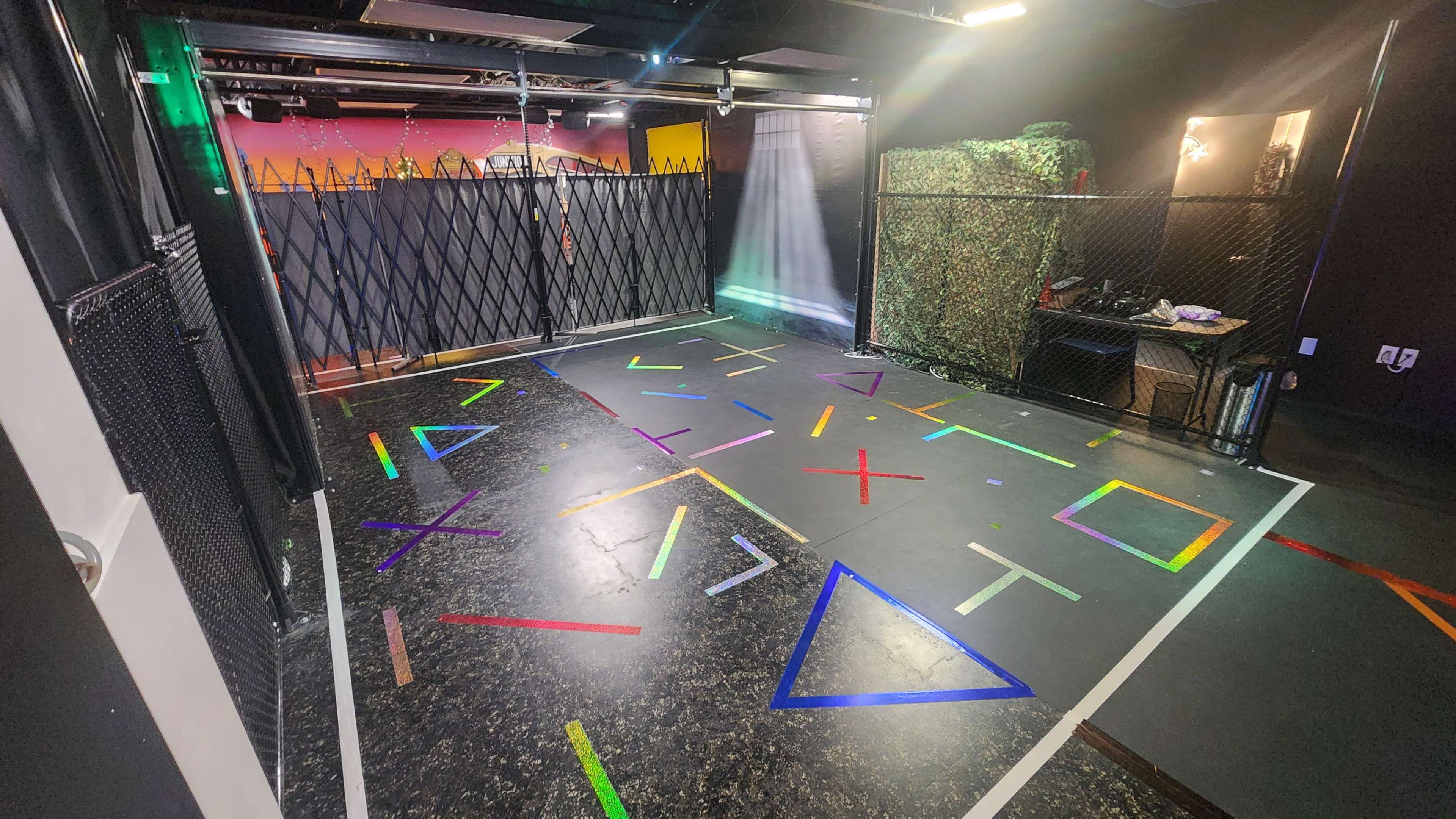 The image shows a room featuring a dark floor with colorful geometric shapes marked on it, surrounded by a wire fence and equipped with various props in the background.