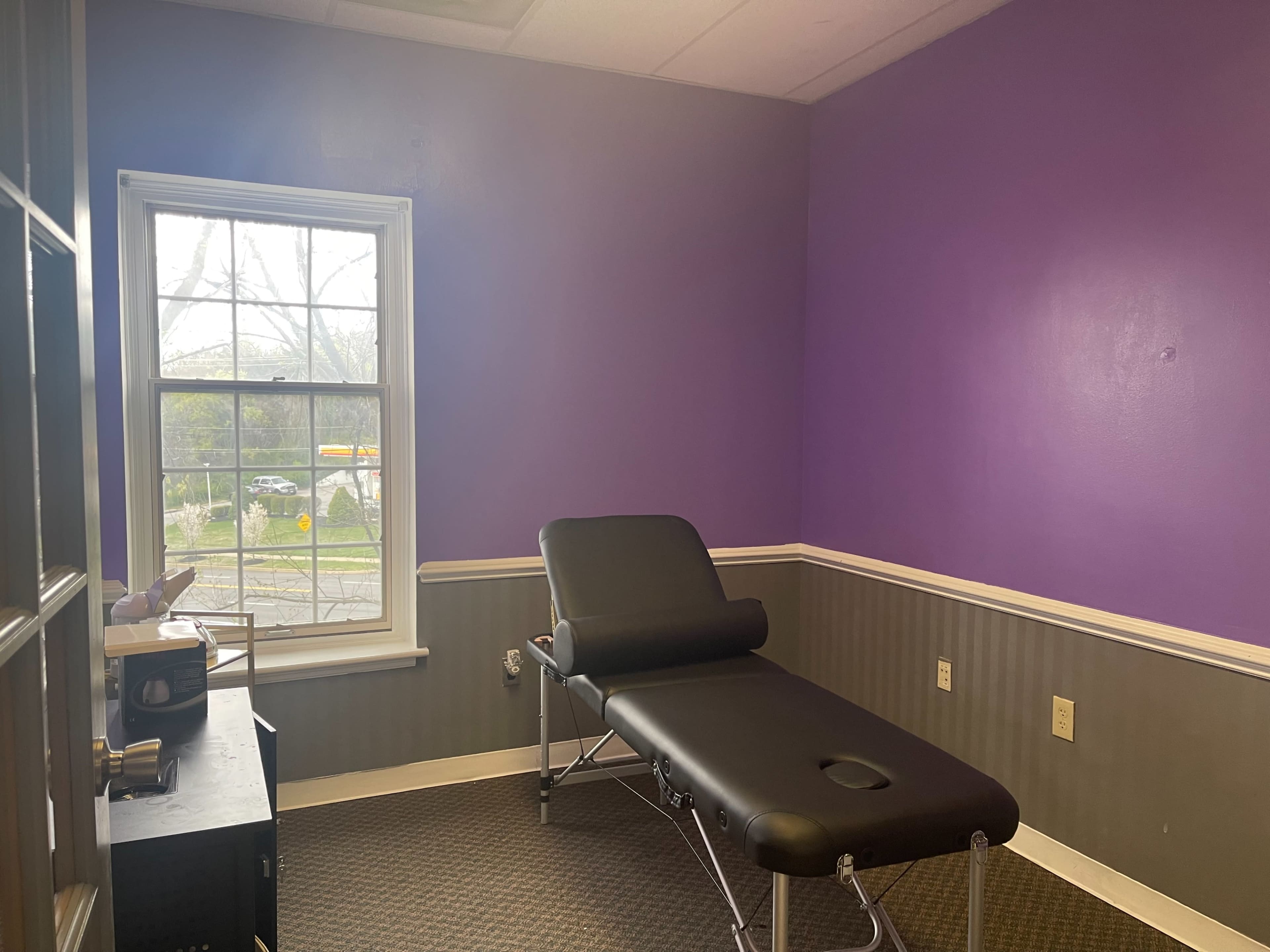 The room features a black treatment table, a small desk, and purple walls with a window showing an outdoor view.