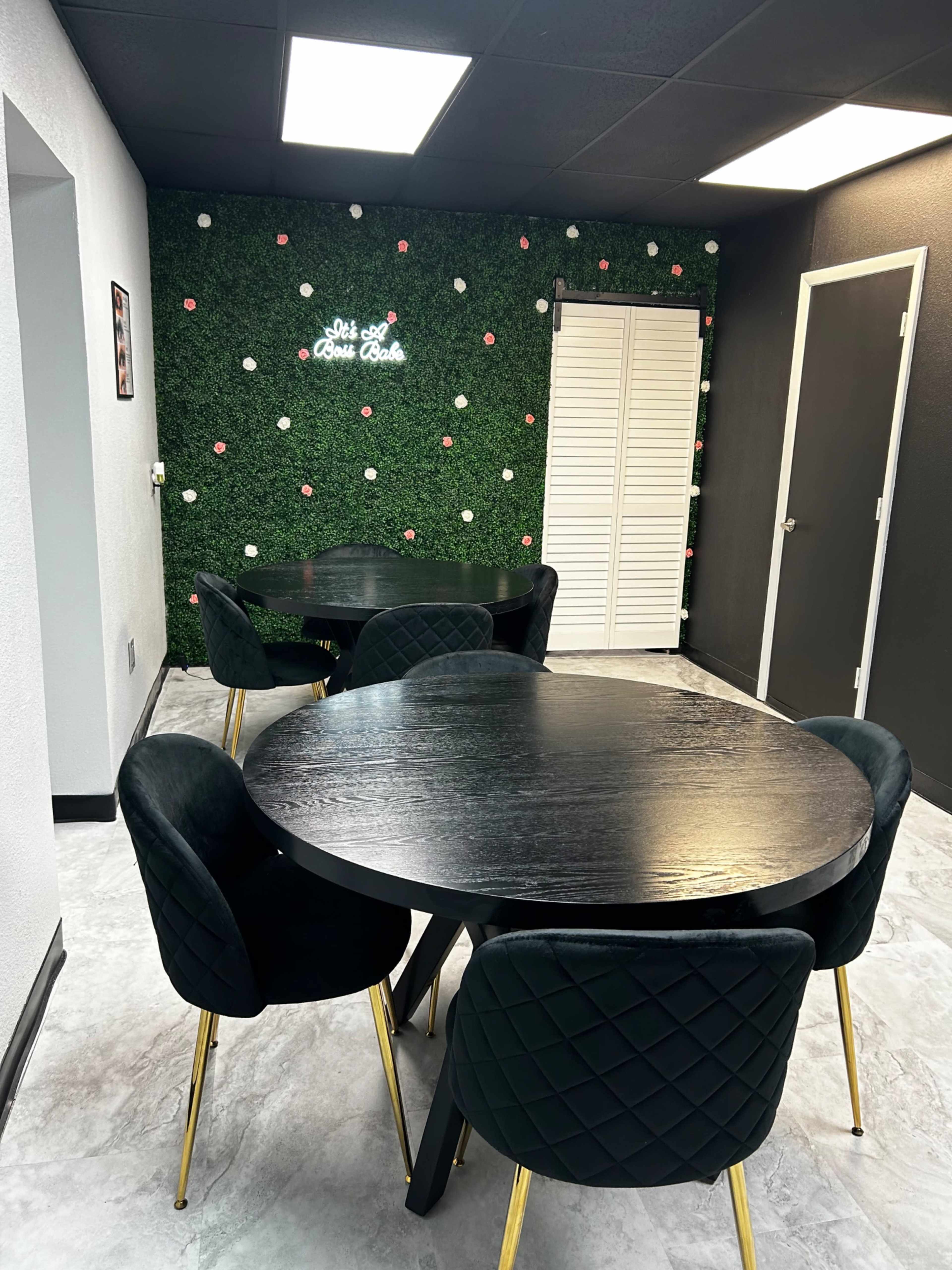The image shows a modern room with dark round tables and black chairs, set against a green wall adorned with white flowers and a neon sign.