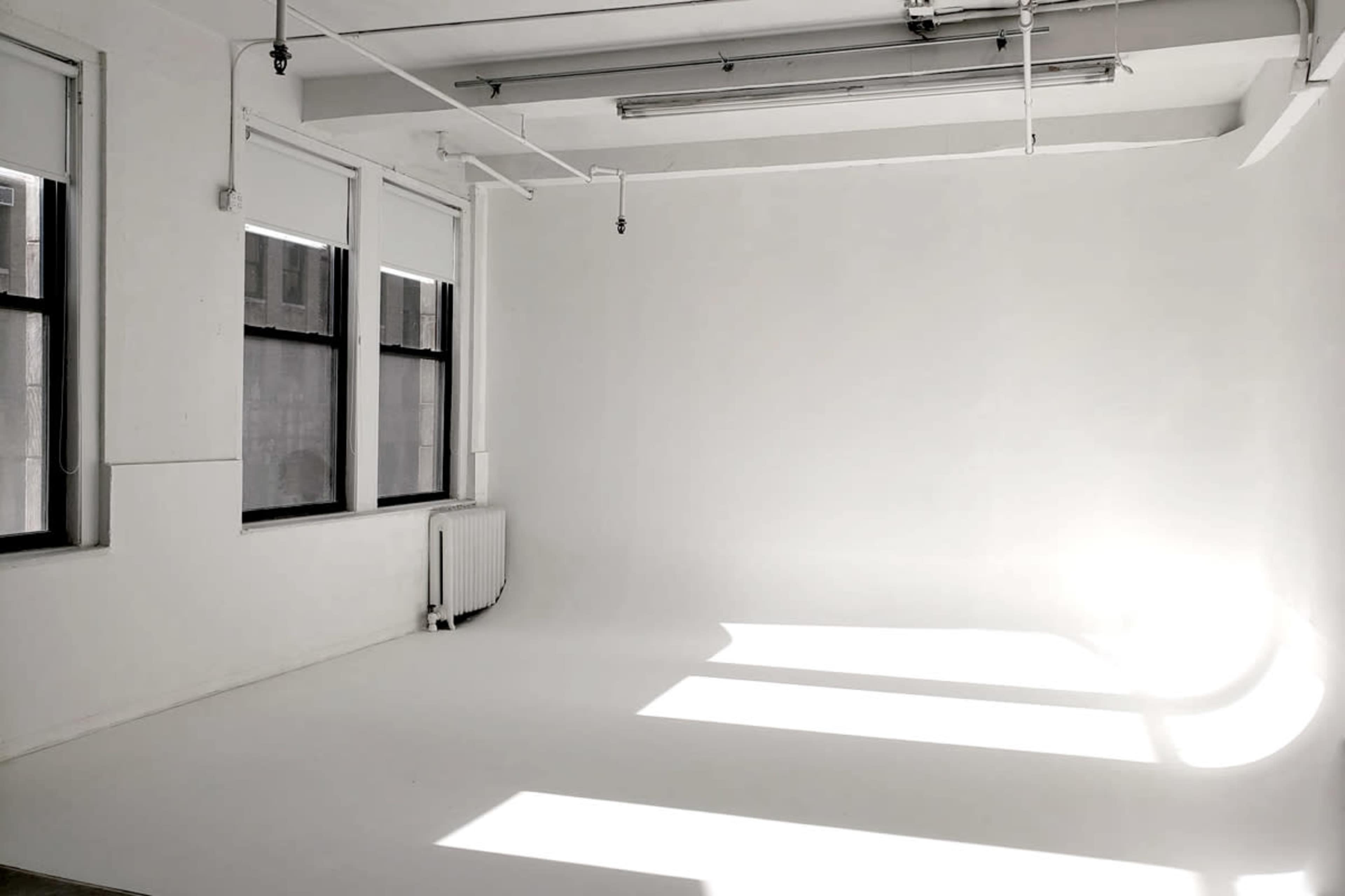 A bright, empty room with white walls and a seamless backdrop, featuring large windows that cast shadows on the floor.