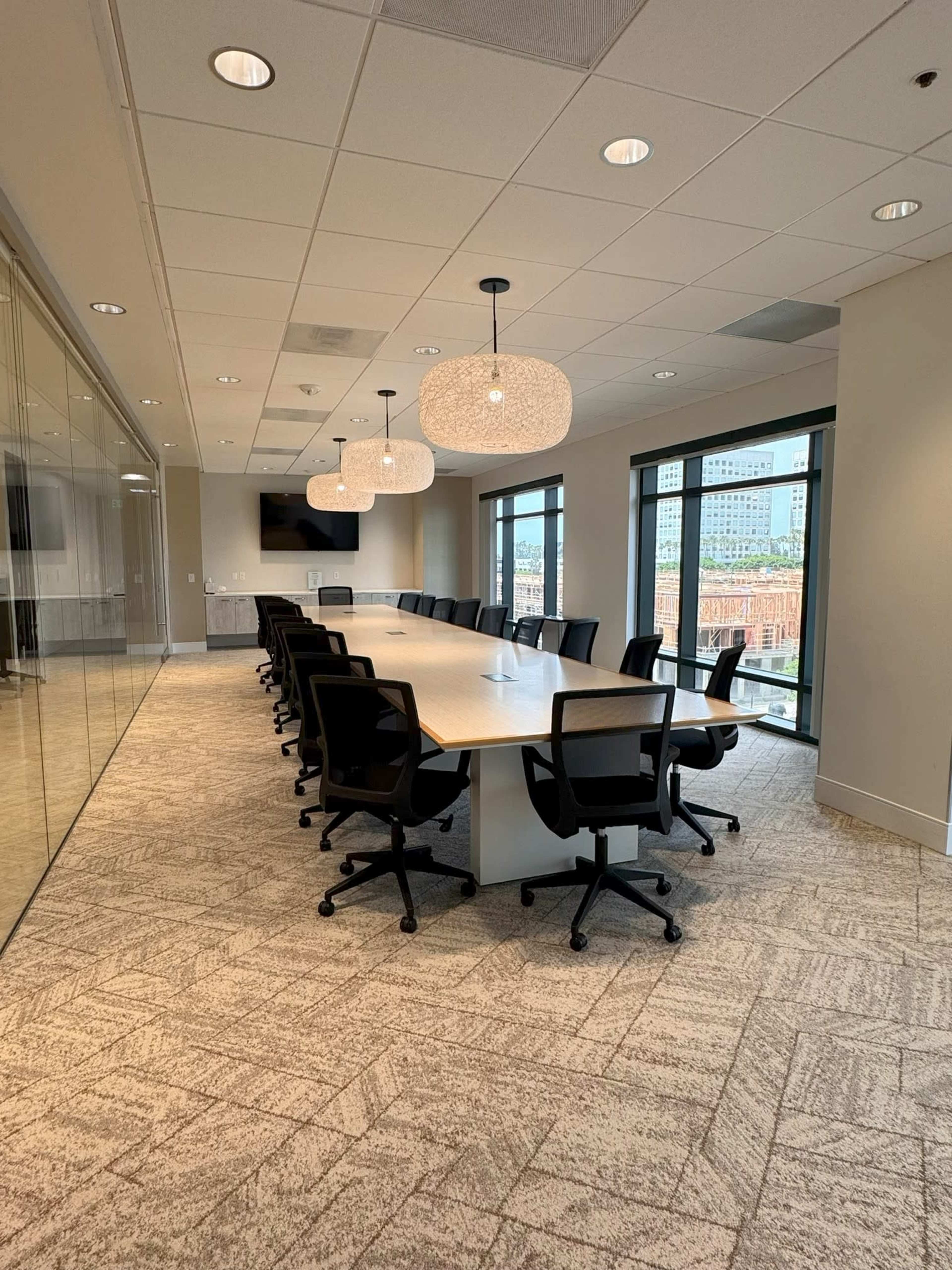 The image shows a modern conference room featuring a long white table surrounded by black swivel chairs, with large windows allowing natural light to fill the space.