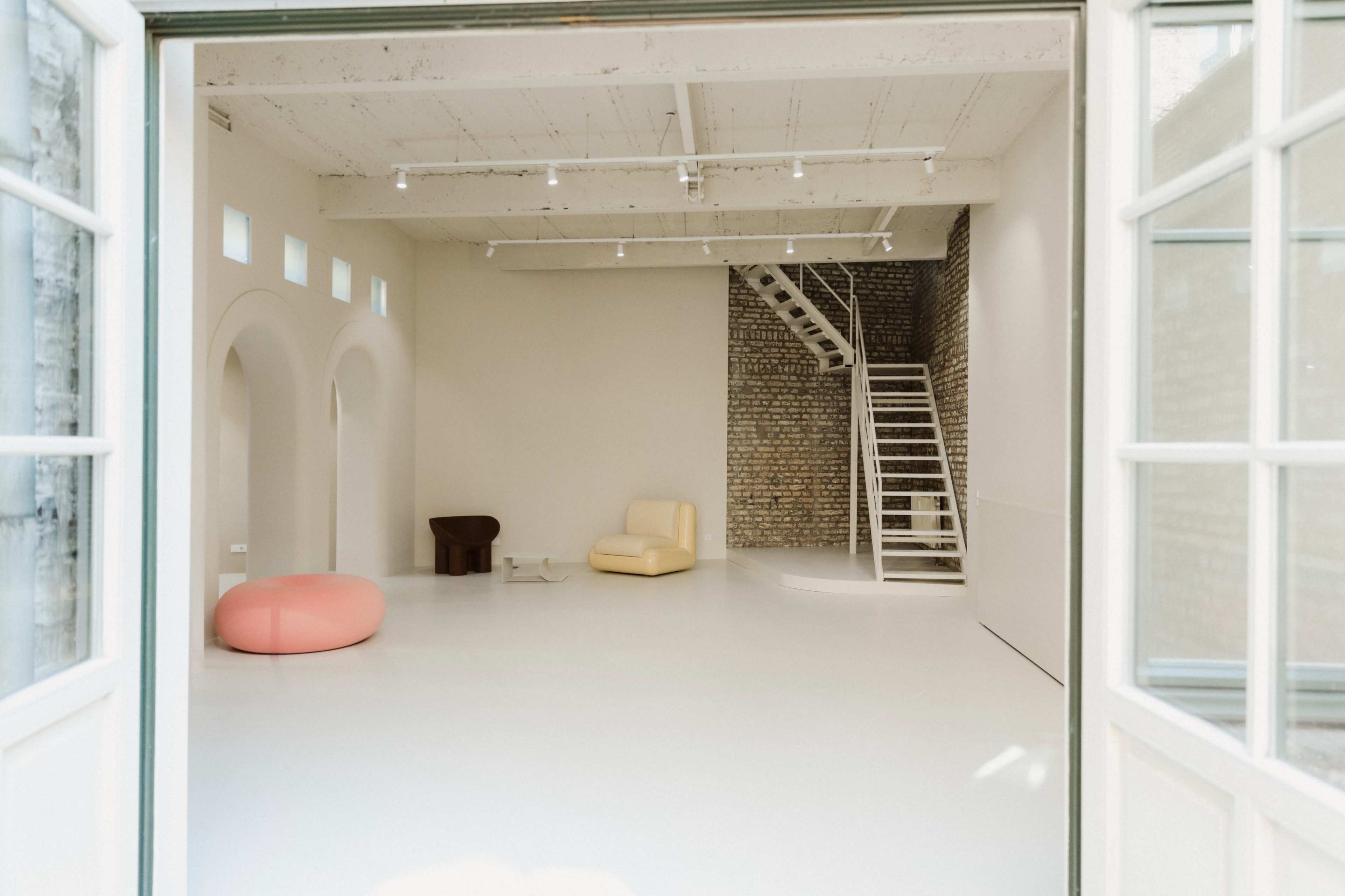 The image shows a spacious, minimalistic room with a white floor and light-colored walls, featuring a few modern furniture pieces, including a pink pouf, a yellow chair, and a dark, rounded table, along with a staircase made of brick in the background.