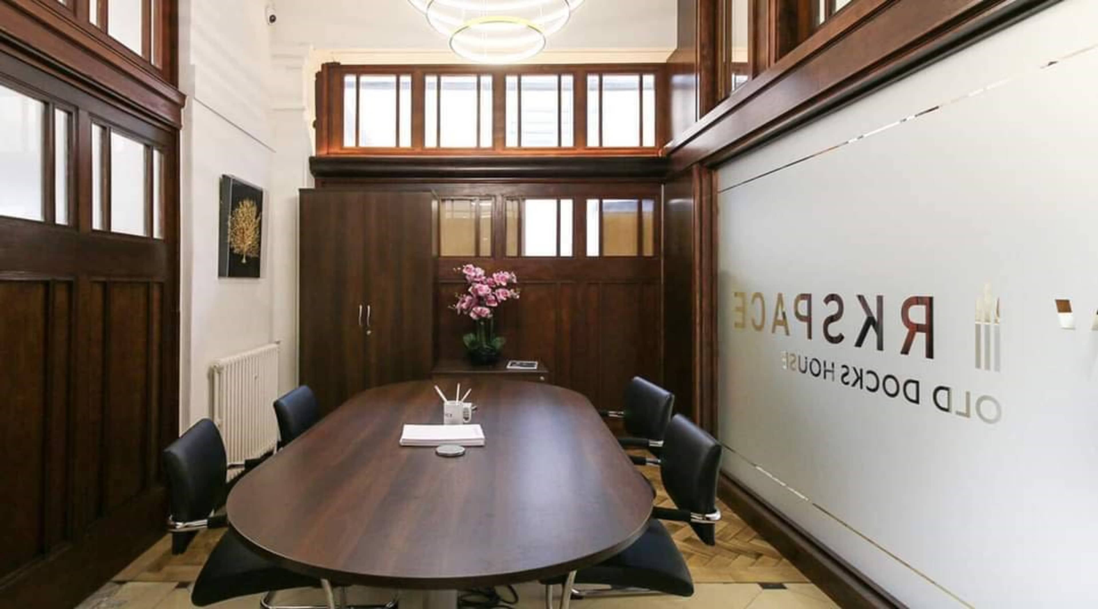 The image shows a modern conference room featuring a wooden table surrounded by black chairs, with a decorative wall and a potted plant.