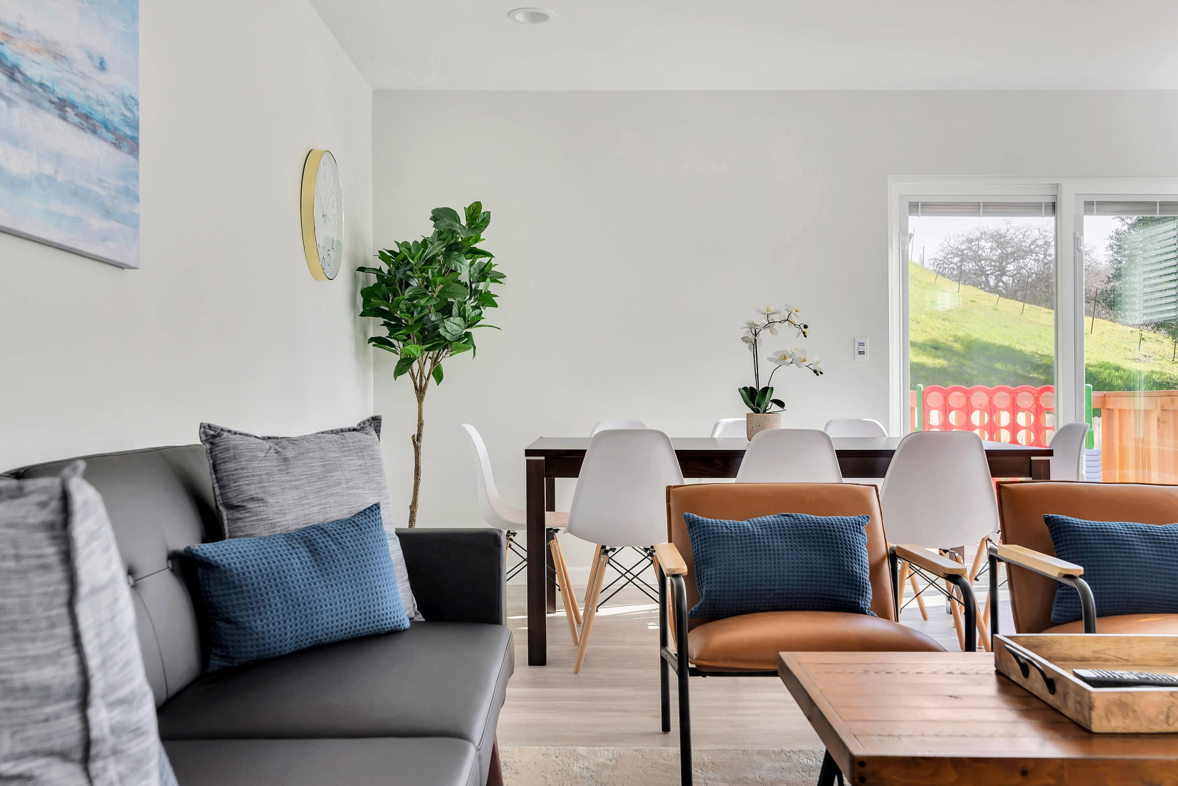 A contemporary living space featuring a gray sofa with blue cushions, a wooden coffee table, and a dining area with a dark table surrounded by white chairs, all positioned near large windows opening to a green hillside.