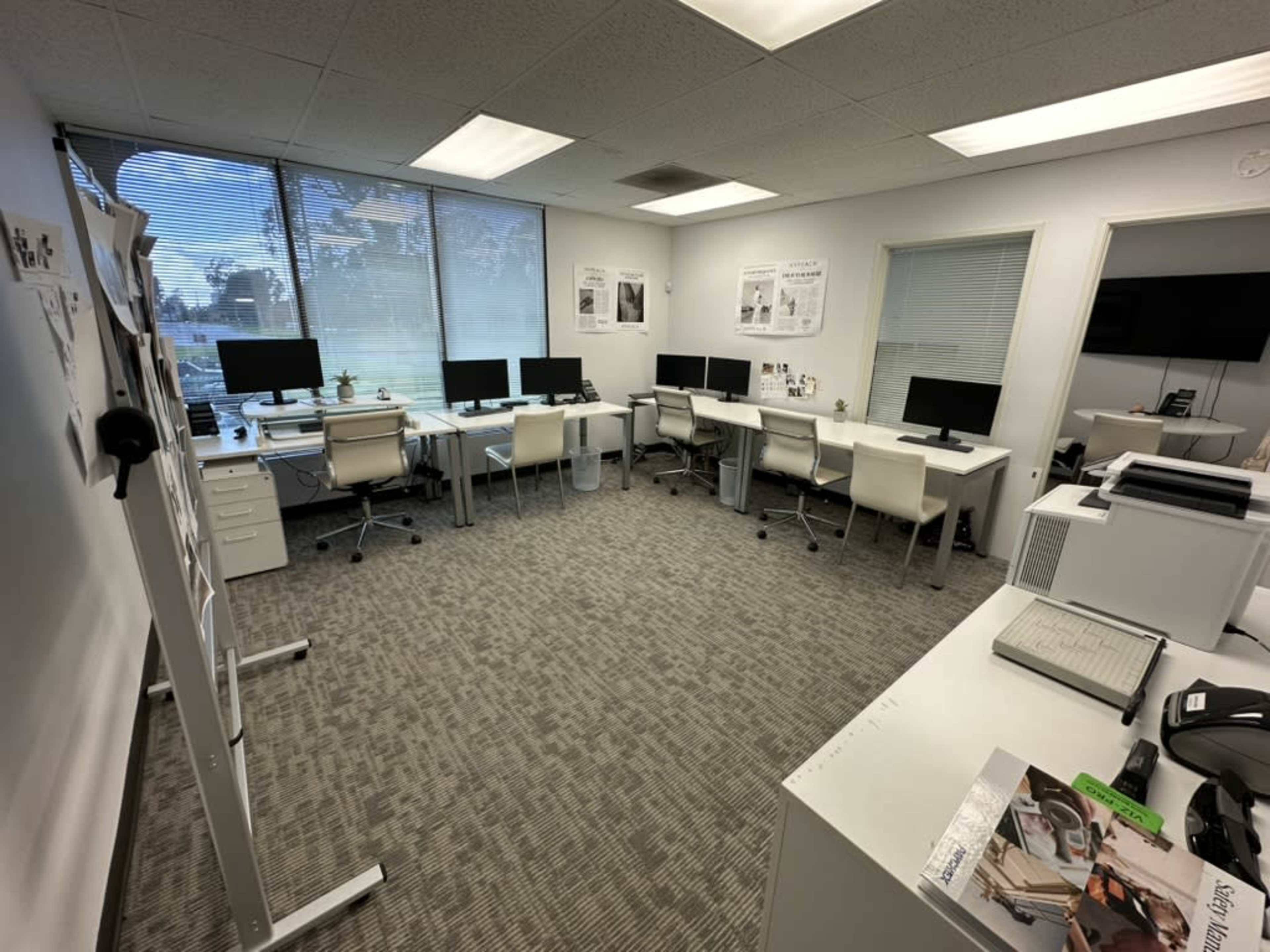 The image shows a modern office space with multiple workstations equipped with computers and ergonomic chairs, along with large windows providing natural light.
