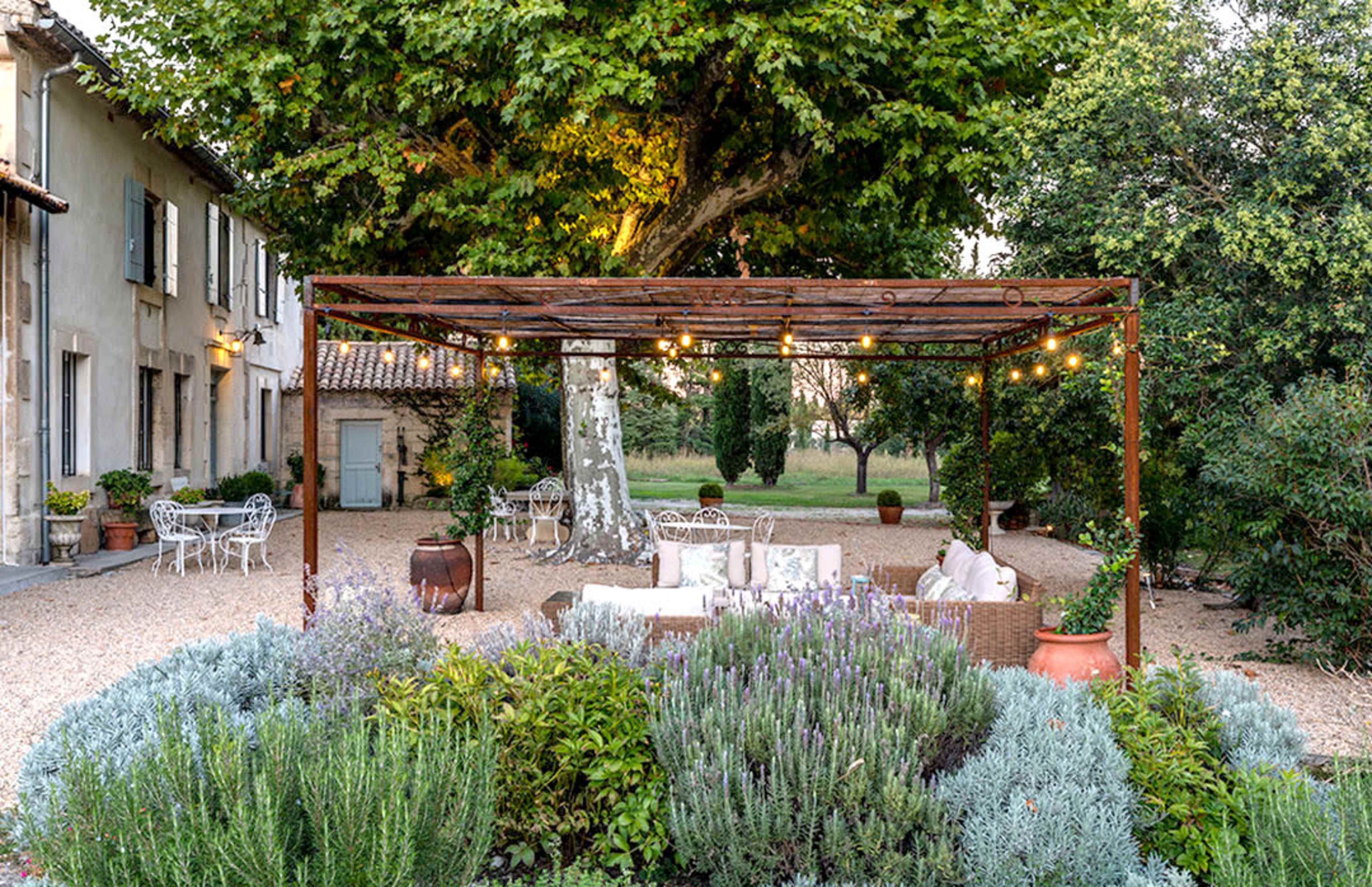 A garden area features a pergola with string lights, surrounded by potted plants and seating arrangements in front of a rustic house.