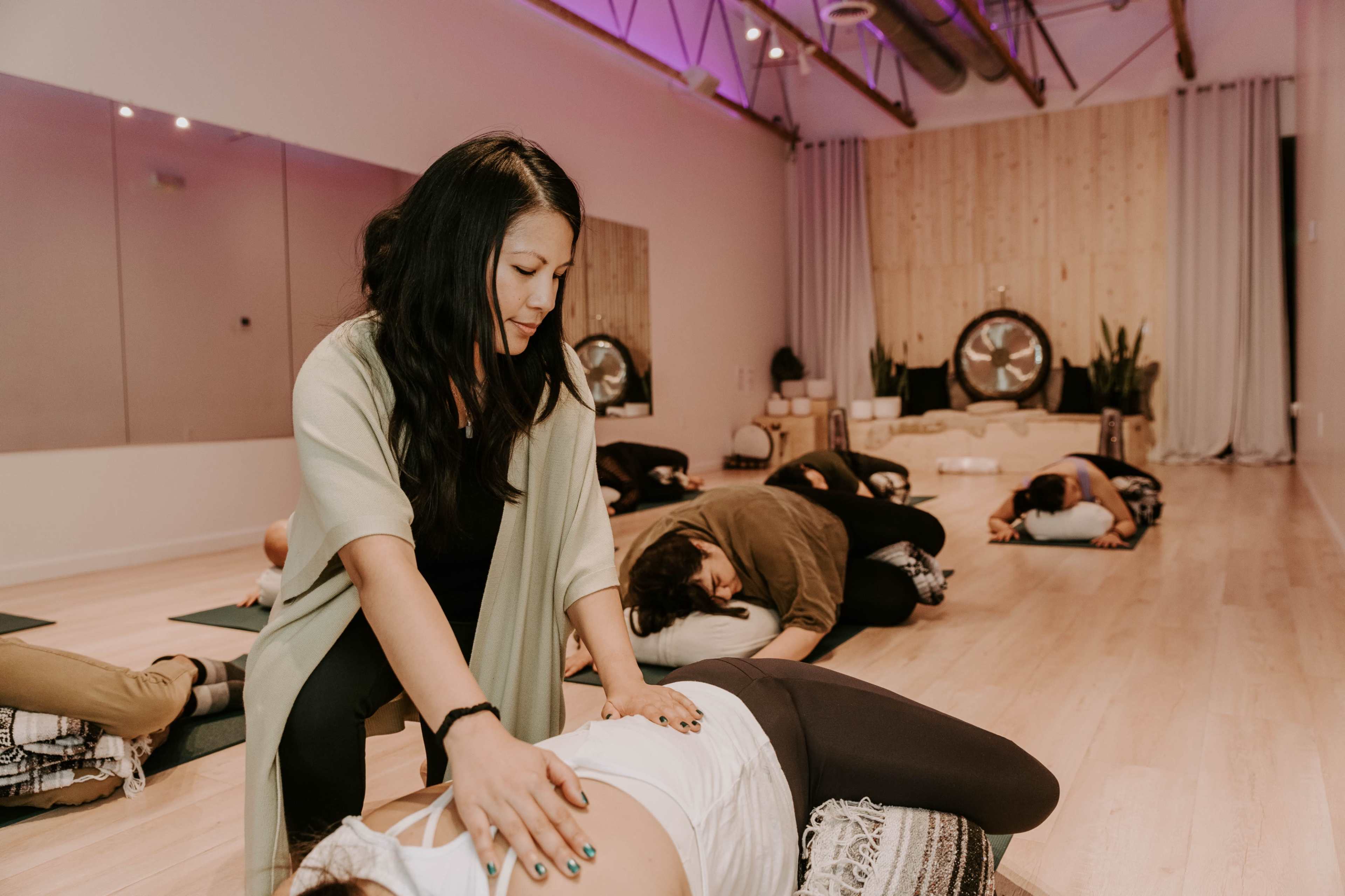 A yoga instructor provides hands-on support to a student during a class in a spacious, well-lit studio.