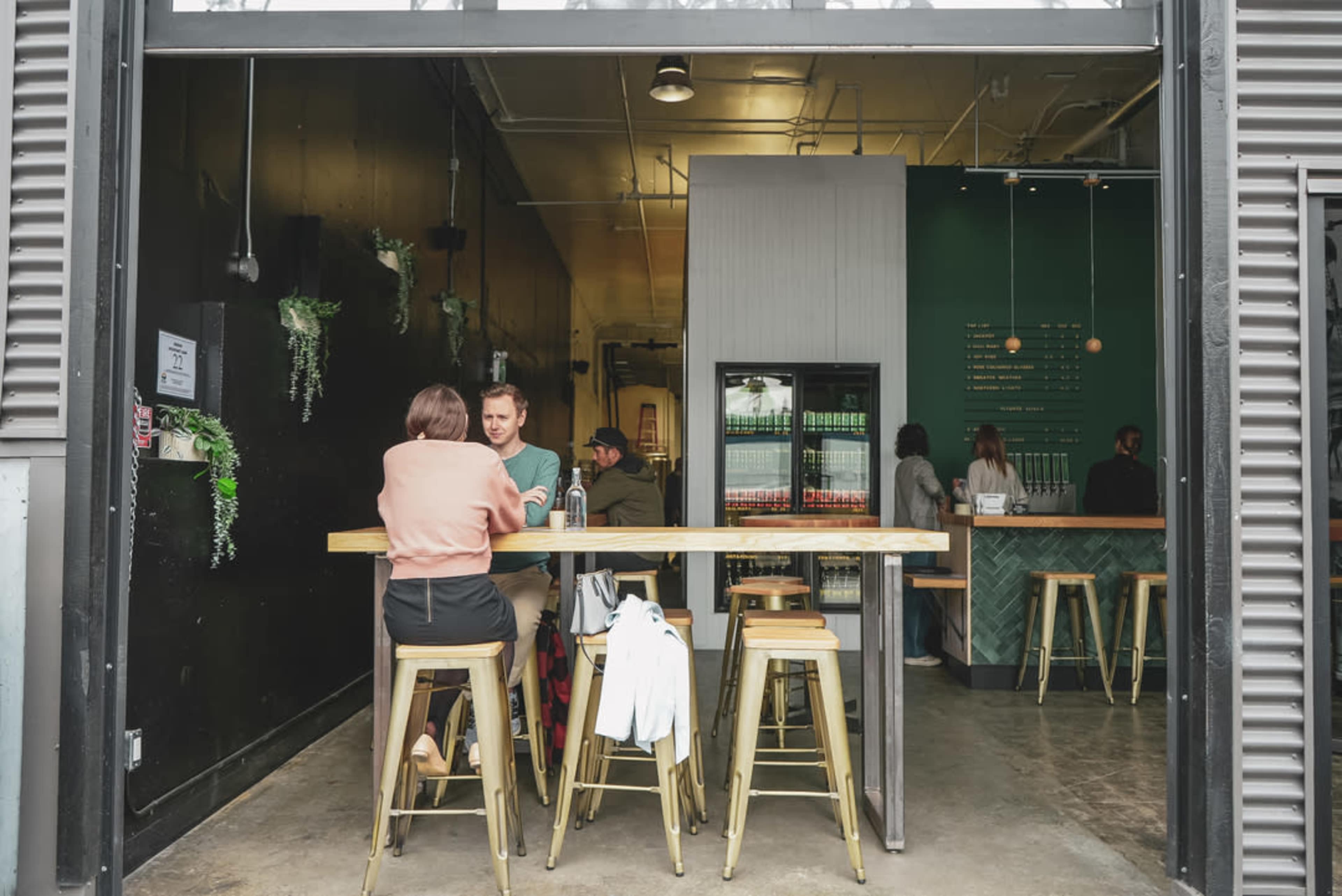 Two people sit at a wooden table inside a cafe, with a beverage cooler and a counter visible in the background.