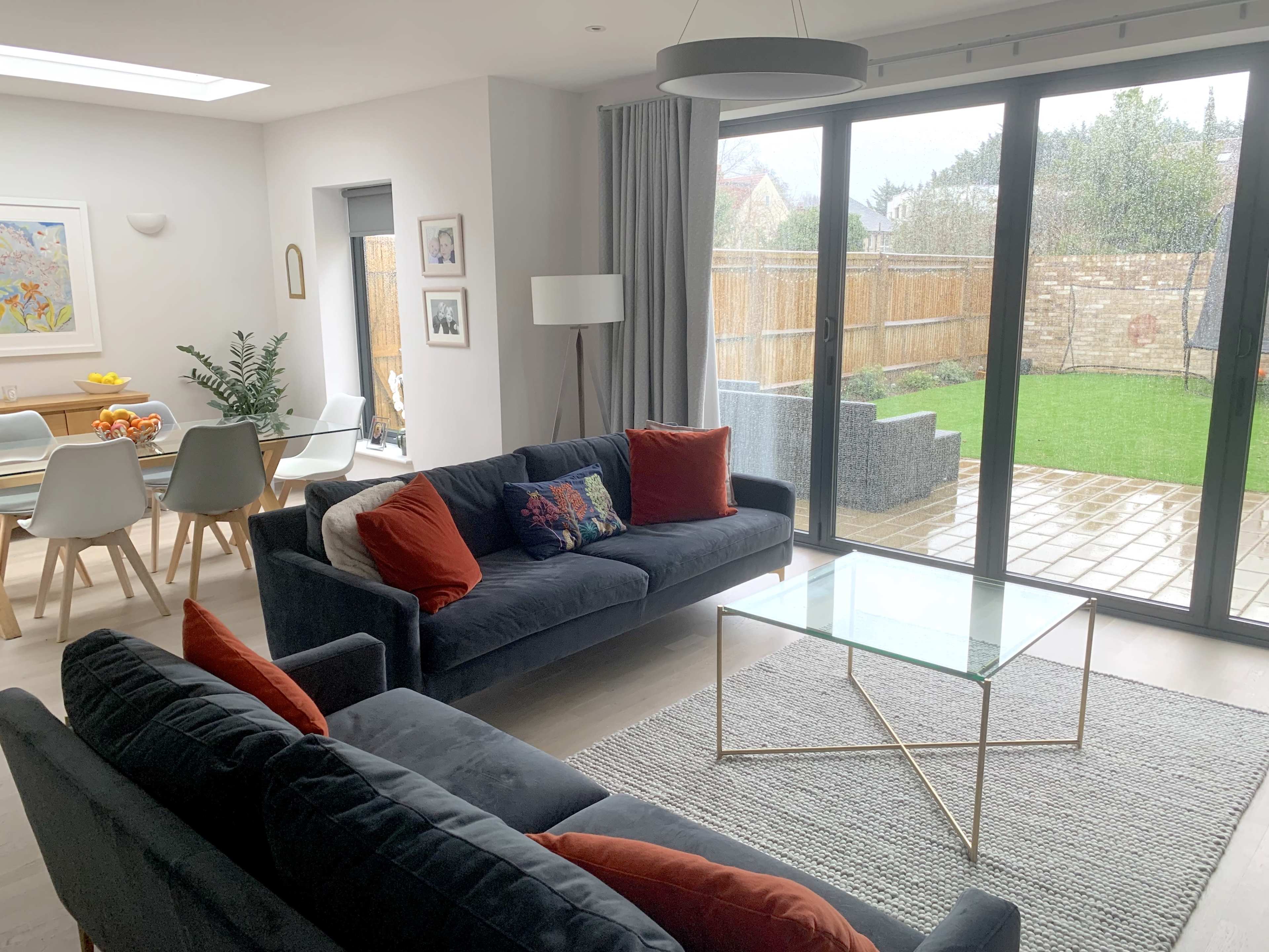 A modern living room with two sofas facing a glass coffee table, large windows overlooking a backyard, and a dining area in the background.