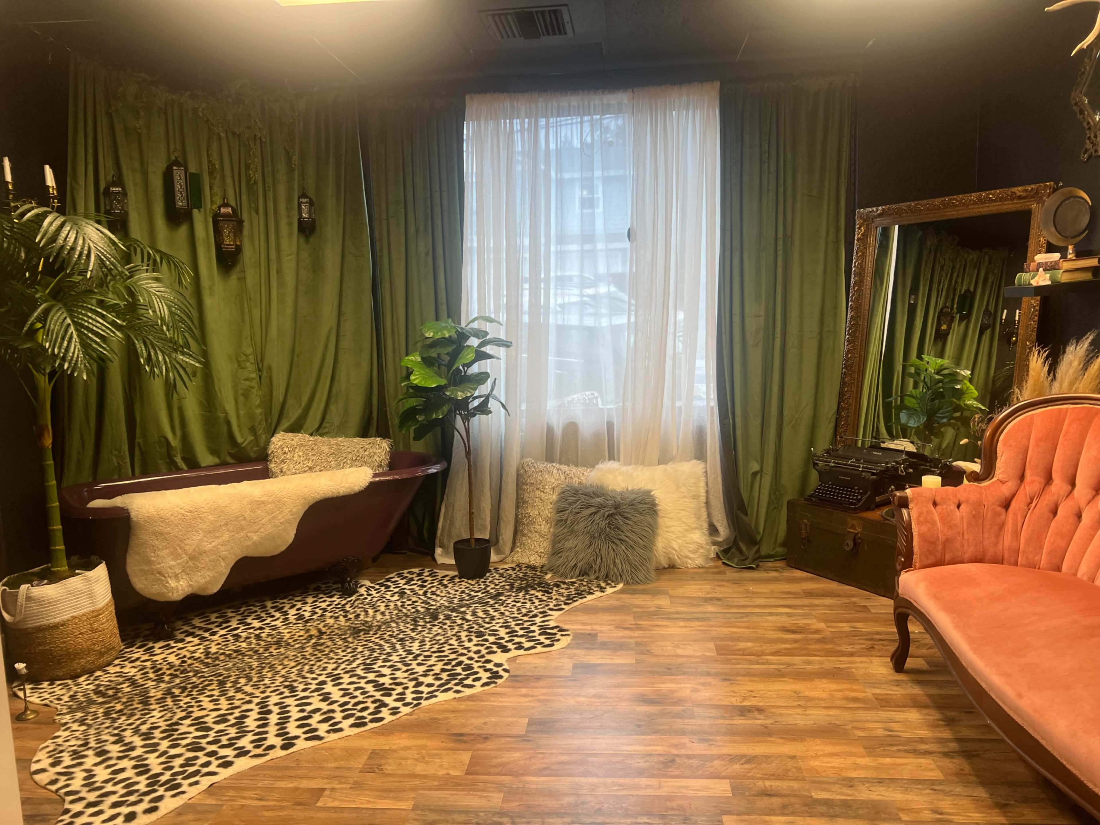 The room features a vintage-style bathtub, a lush green curtain, an orange velvet couch, and a patterned rug on a wooden floor.