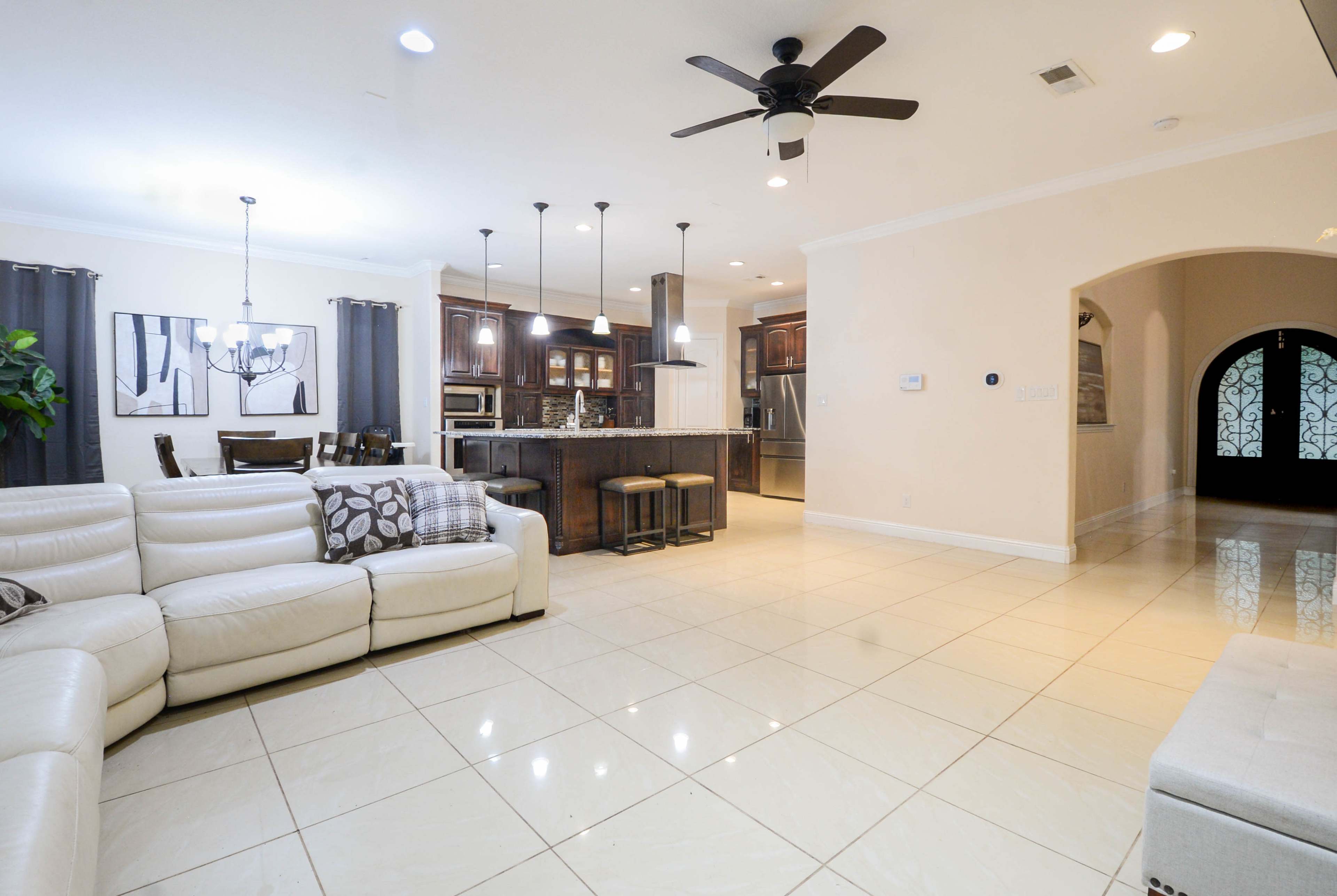 The image shows a modern, open-concept living room with a light-colored leather sectional sofa, hardwood dining furniture, and a kitchen area with dark cabinetry and pendant lighting.