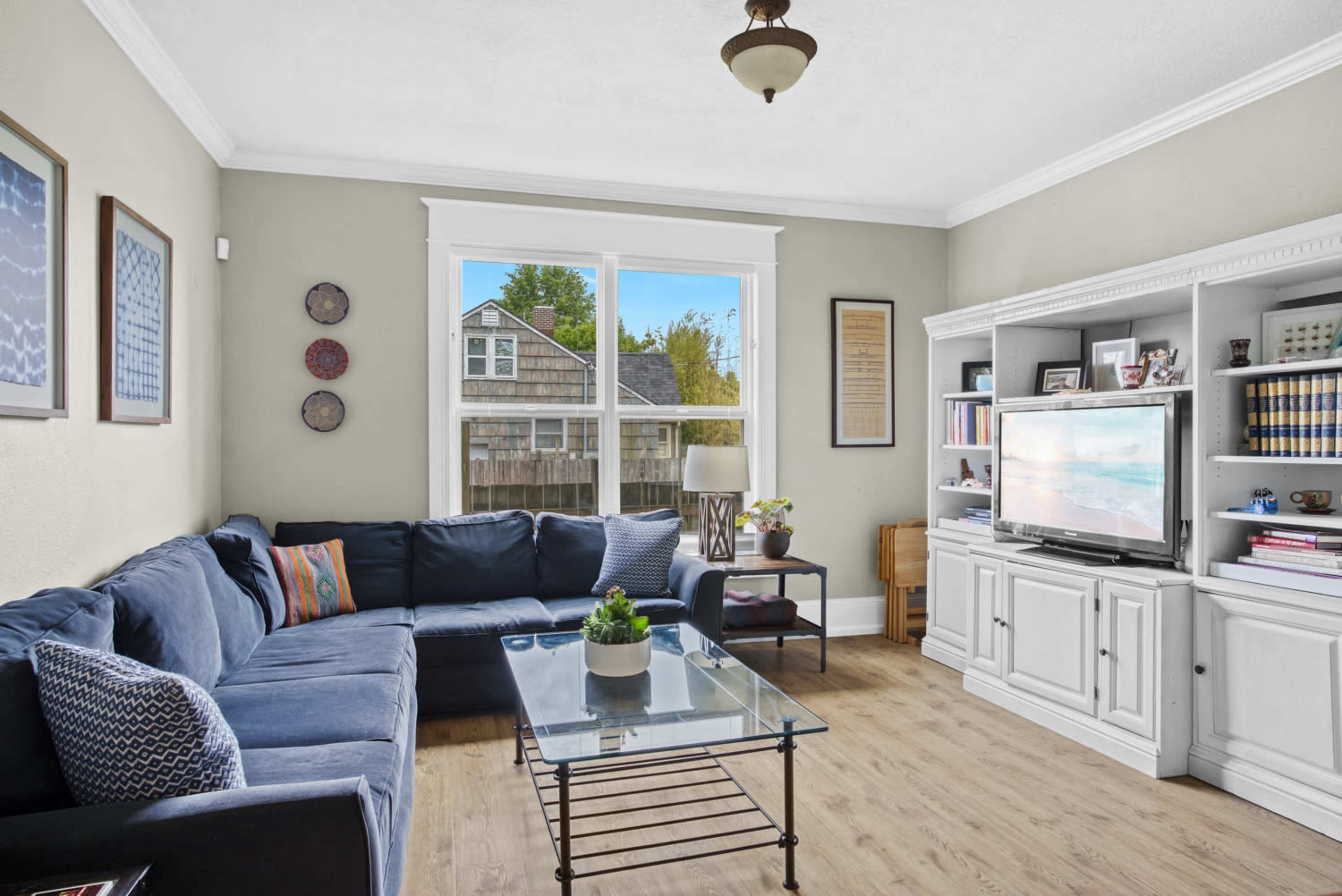 The image shows a living room with a blue sectional sofa, a glass coffee table, and a television on a white cabinet, with a large window revealing a view of the outdoors.