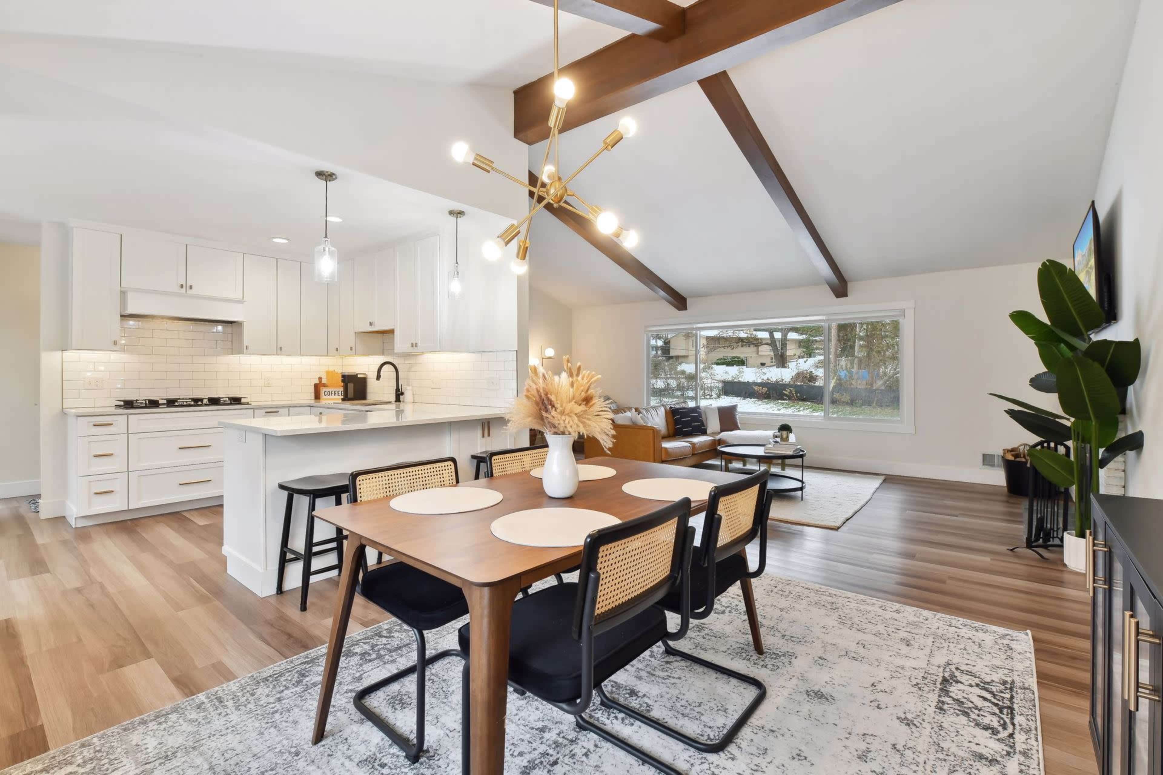 A modern living space features a dining area with a table and chairs, a kitchen with white cabinetry, and a cozy sitting area illuminated by natural light through large windows.