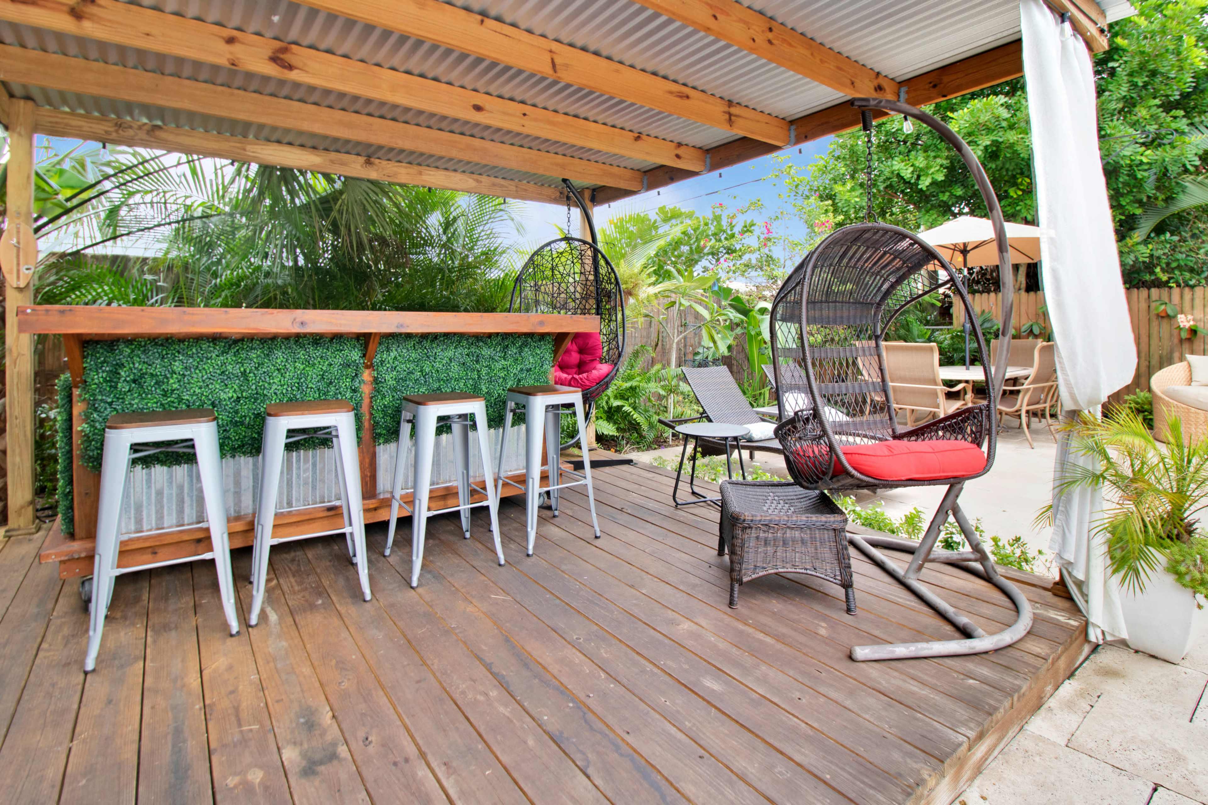 The image shows a wooden deck area featuring a bar with metal stools, a hanging chair, and surrounding greenery.