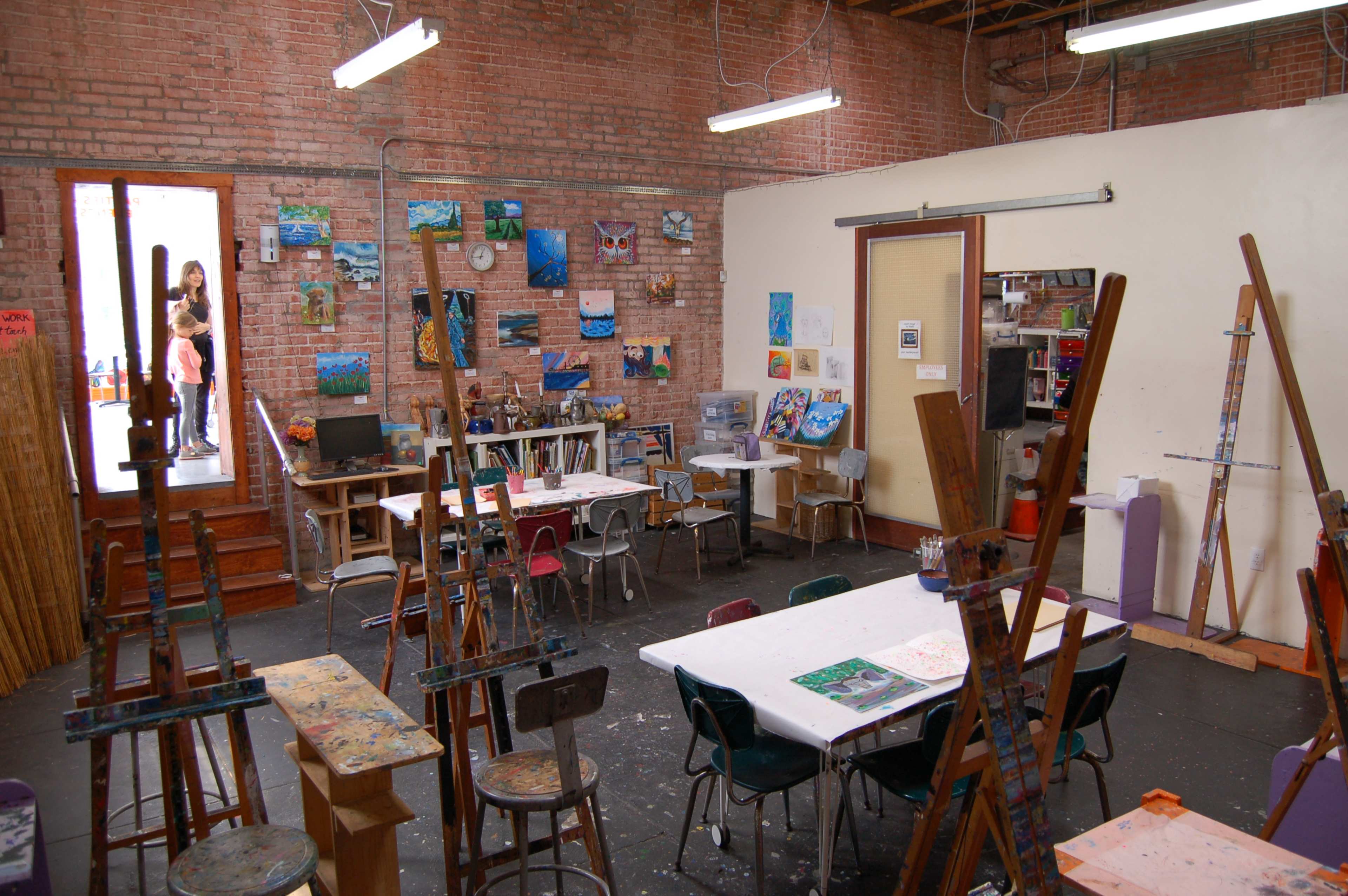 An art studio features easels and tables surrounded by paintings on the walls, with an entrance visible in the background.