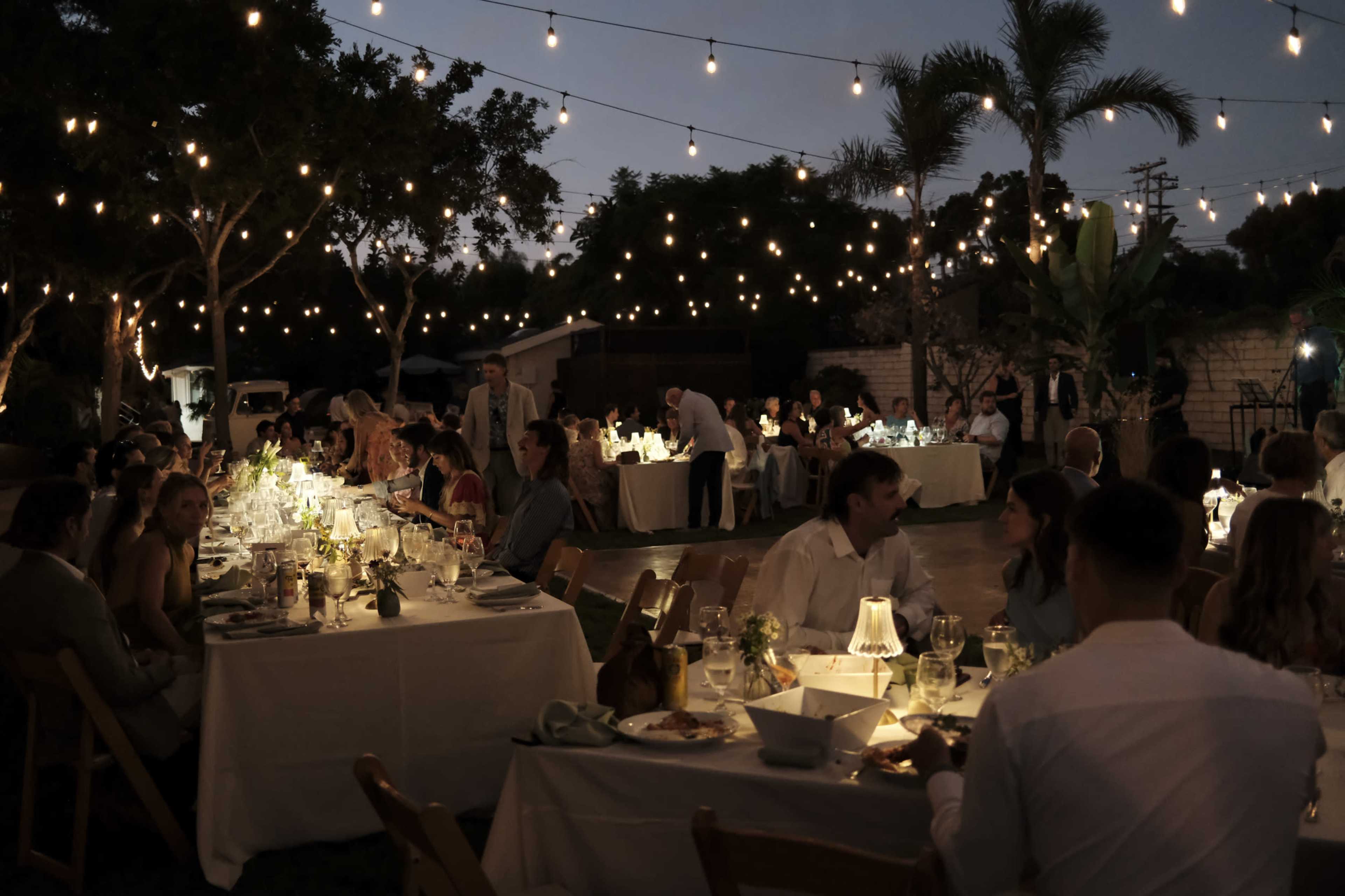 A garden party is set at dusk, featuring tables elegantly arranged with decorations and illuminated by string lights.