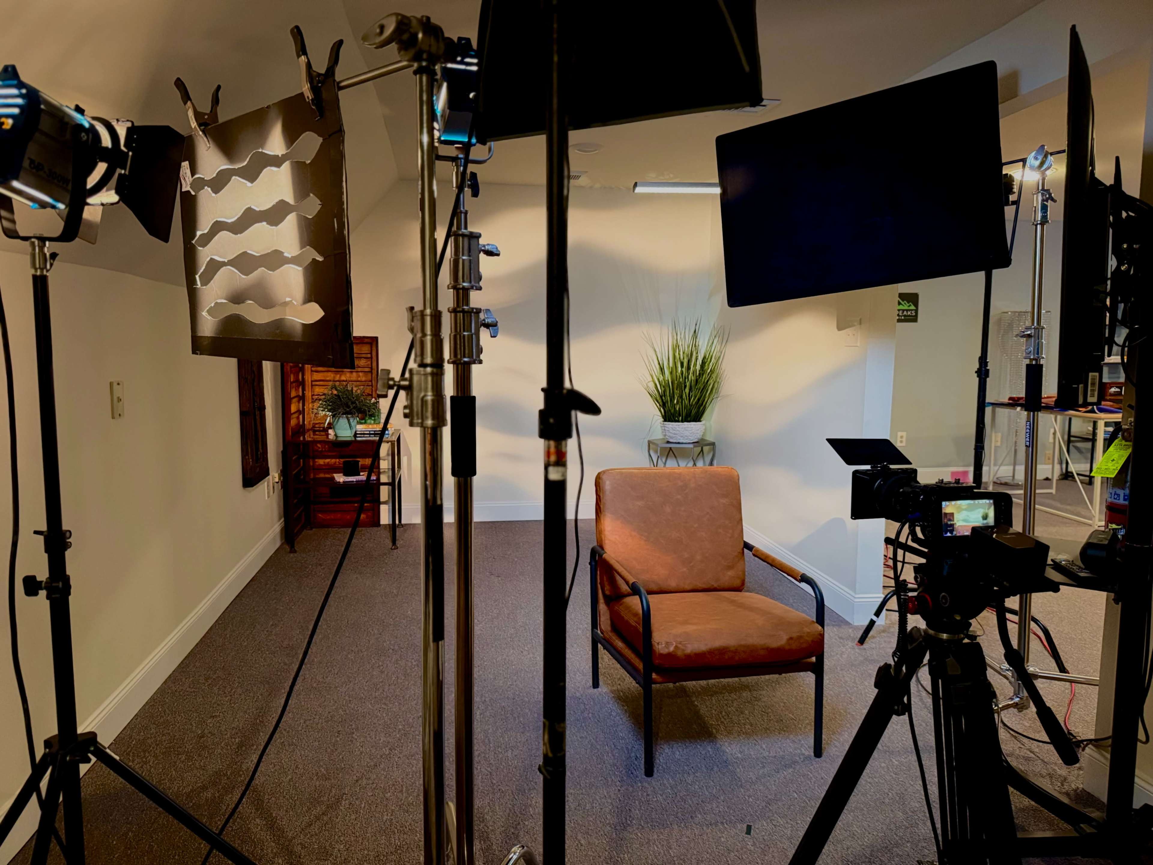 The image shows a video recording setup featuring a brown chair, lights, and cameras arranged around a small, plant-filled backdrop in a room with gray carpeting.