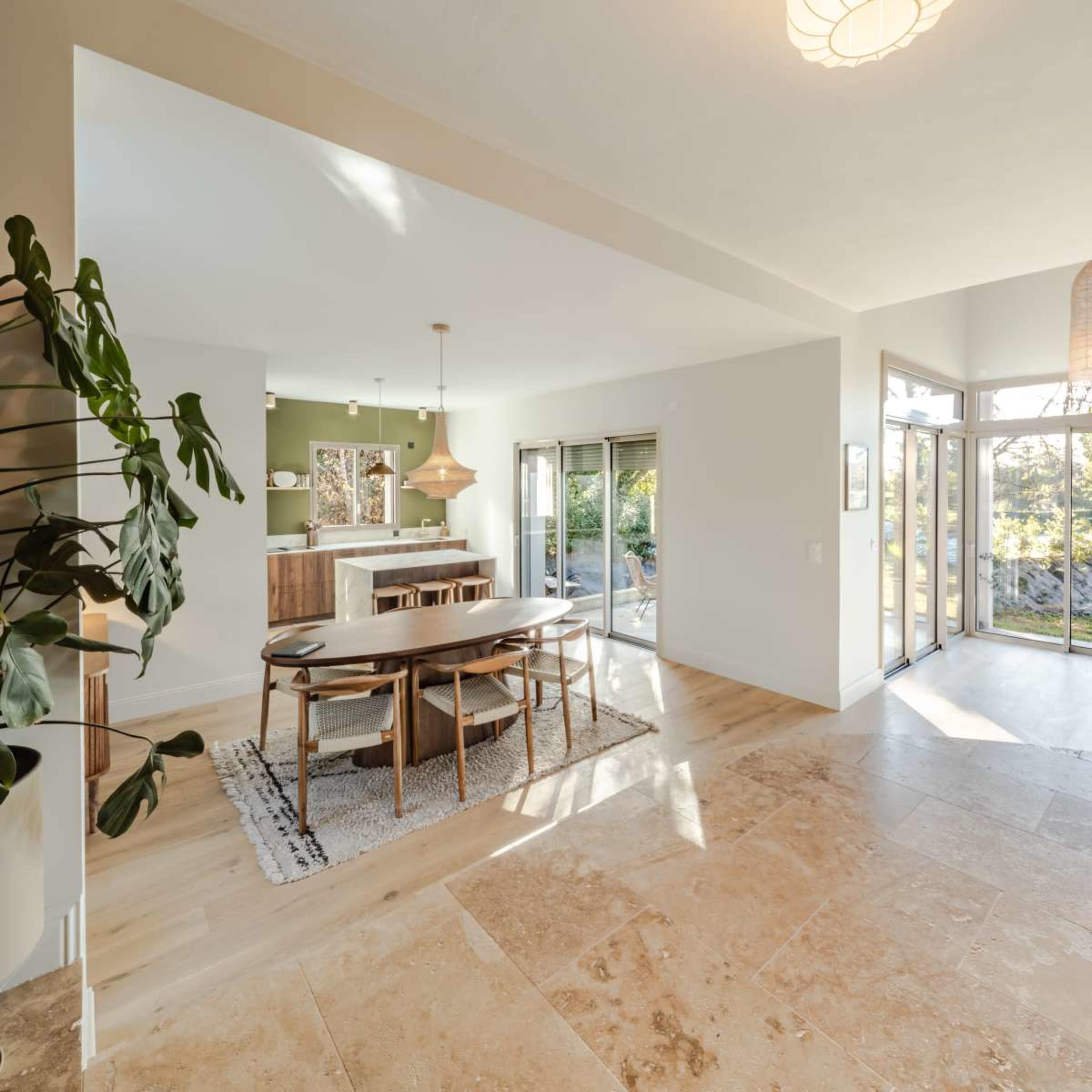 The image shows a bright, open living space featuring a dining area with a wooden table and chairs, connected to a kitchen with modern fixtures and large sliding glass doors leading to an outdoor area.