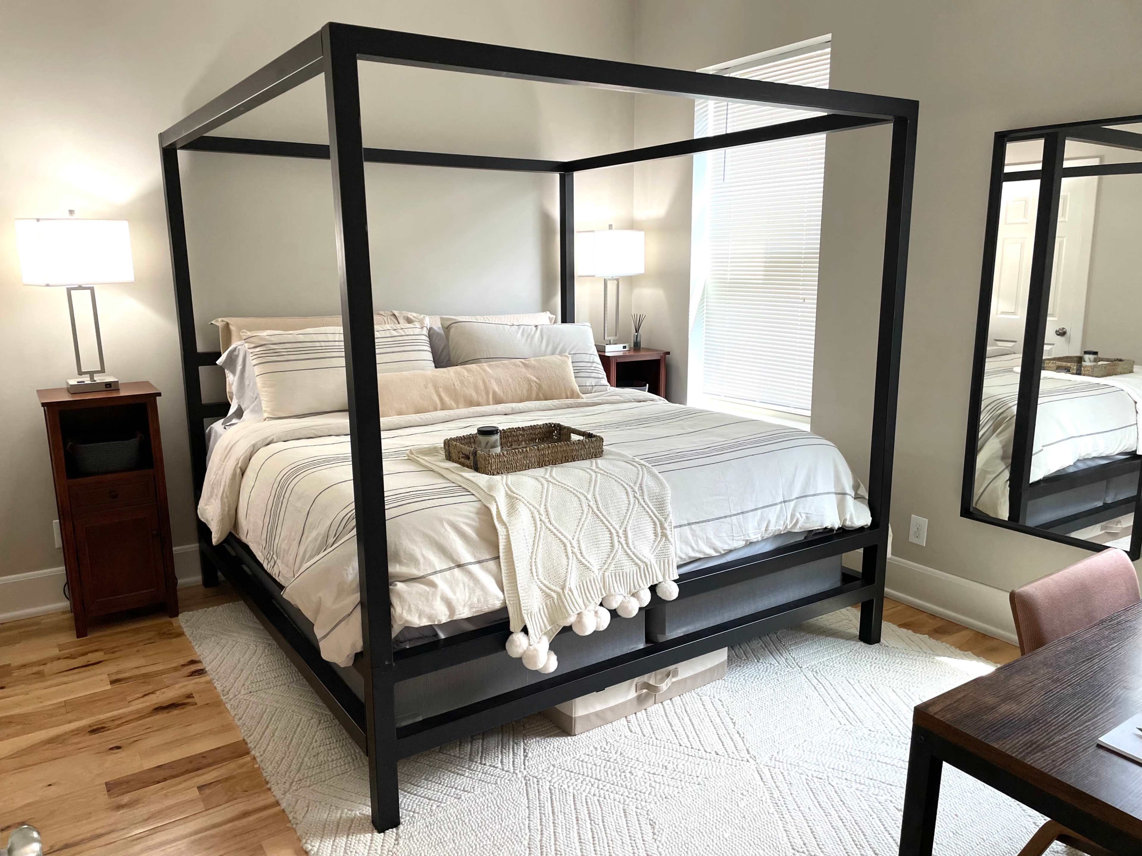 A modern bedroom featuring a four-poster bed with a light-colored comforter, two bedside lamps, and a mirror reflecting a desk area.