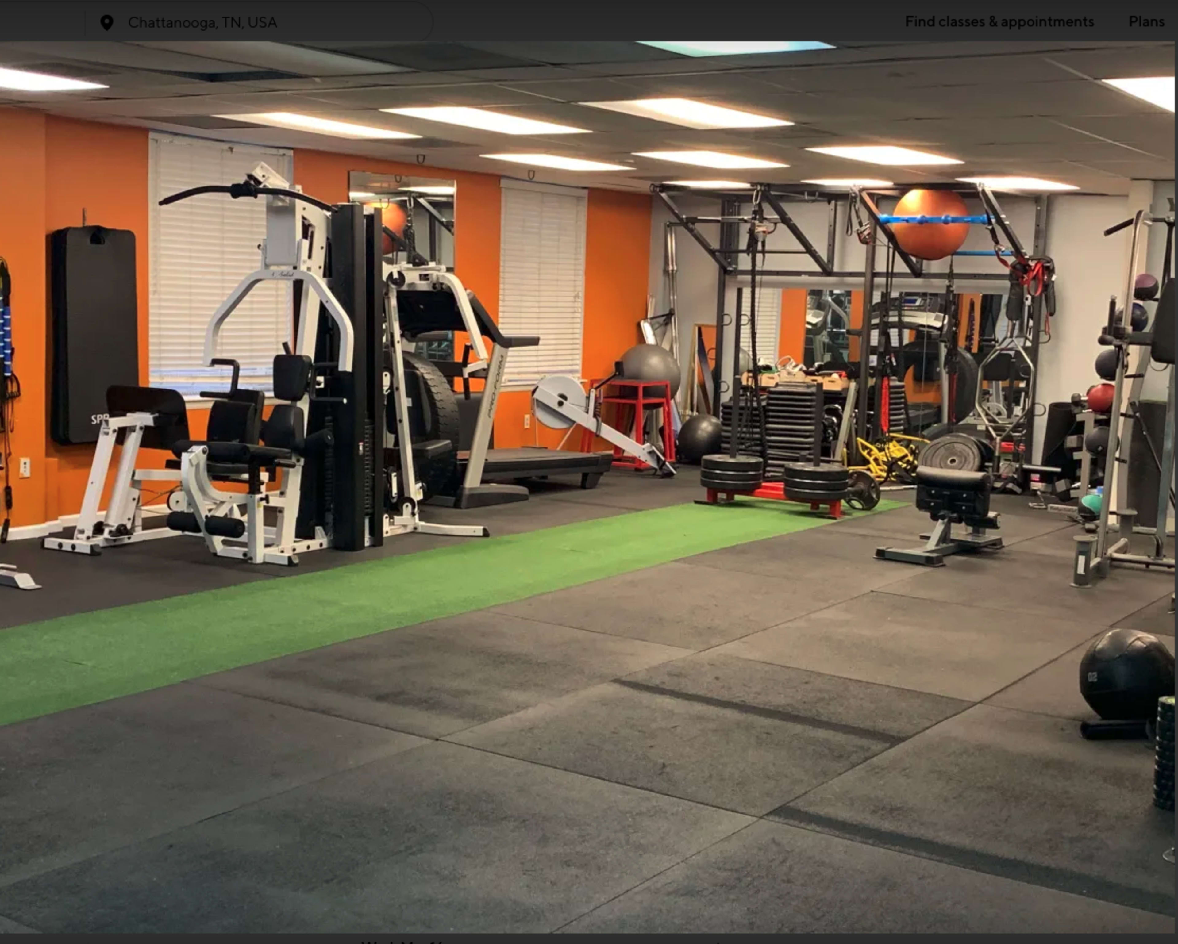A fitness gym equipped with various exercise machines and weights, featuring a green turf area and black rubber flooring.