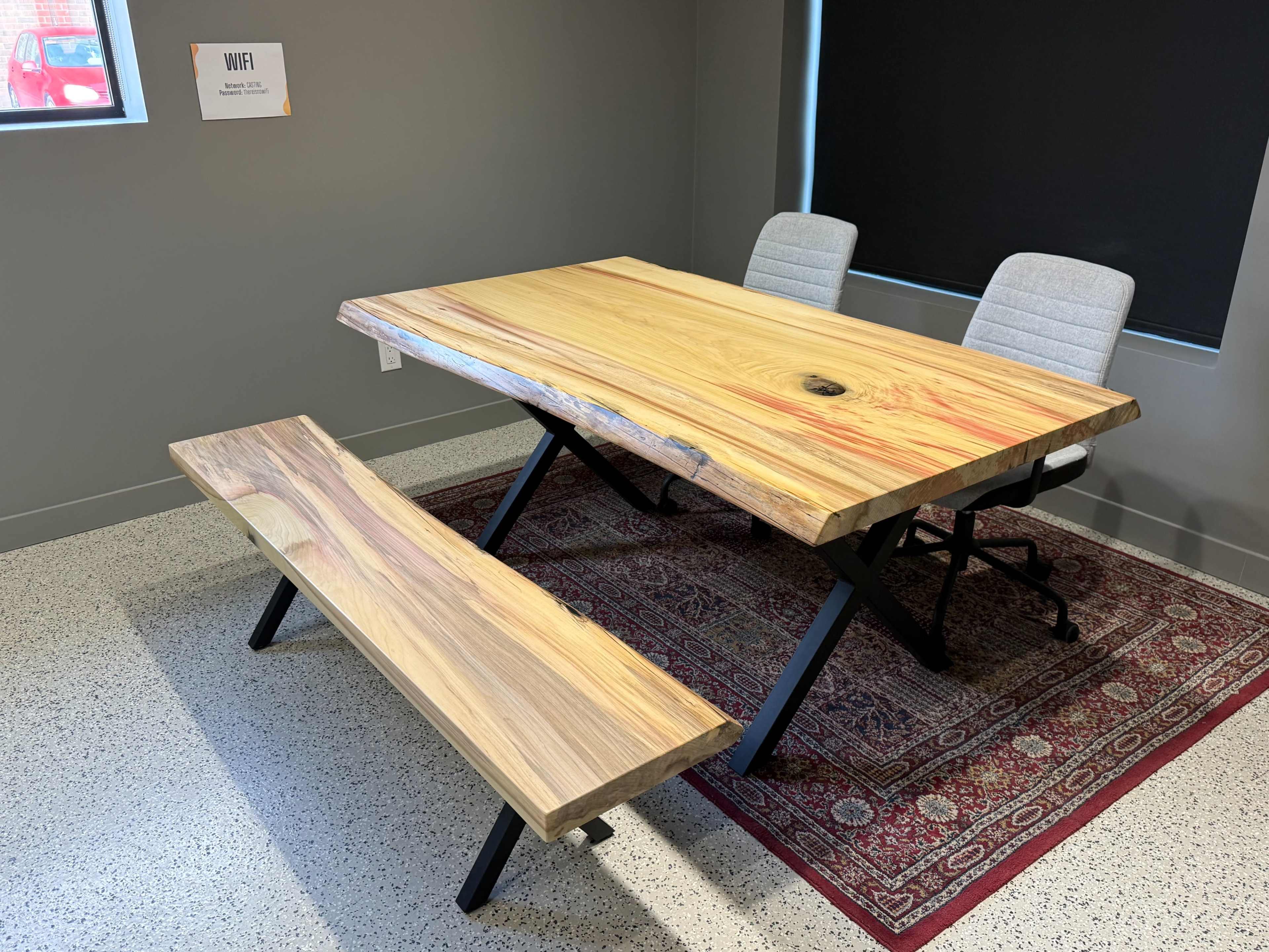 The image shows a large wooden table with a natural edge, accompanied by a matching bench and two gray office chairs, all set on a patterned rug in a well-lit room with a black window covering.