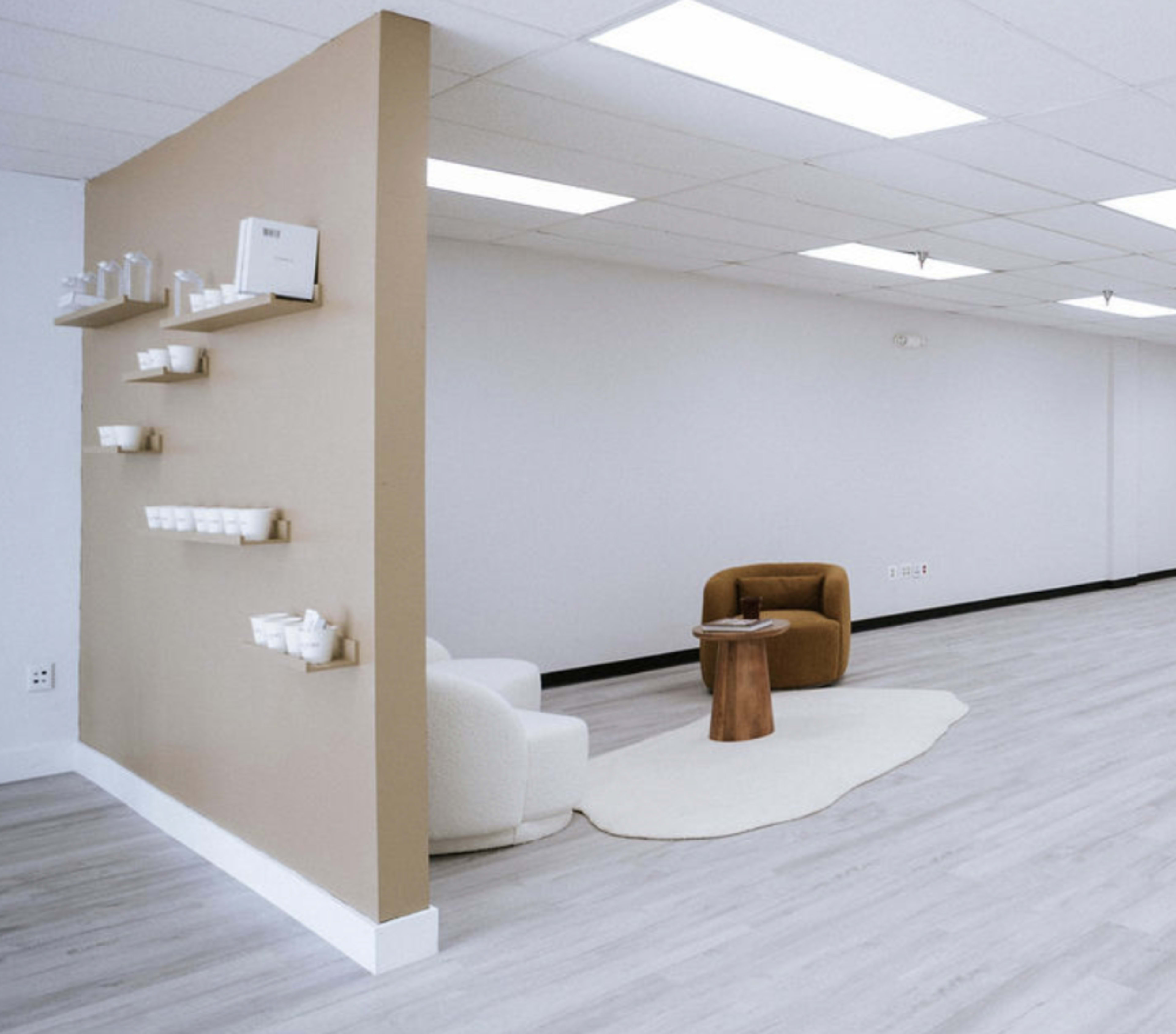 The image shows a minimalist interior featuring a light-colored floor, a beige accent wall with shelves, and two modern chairs arranged around a small wooden table on a rug.