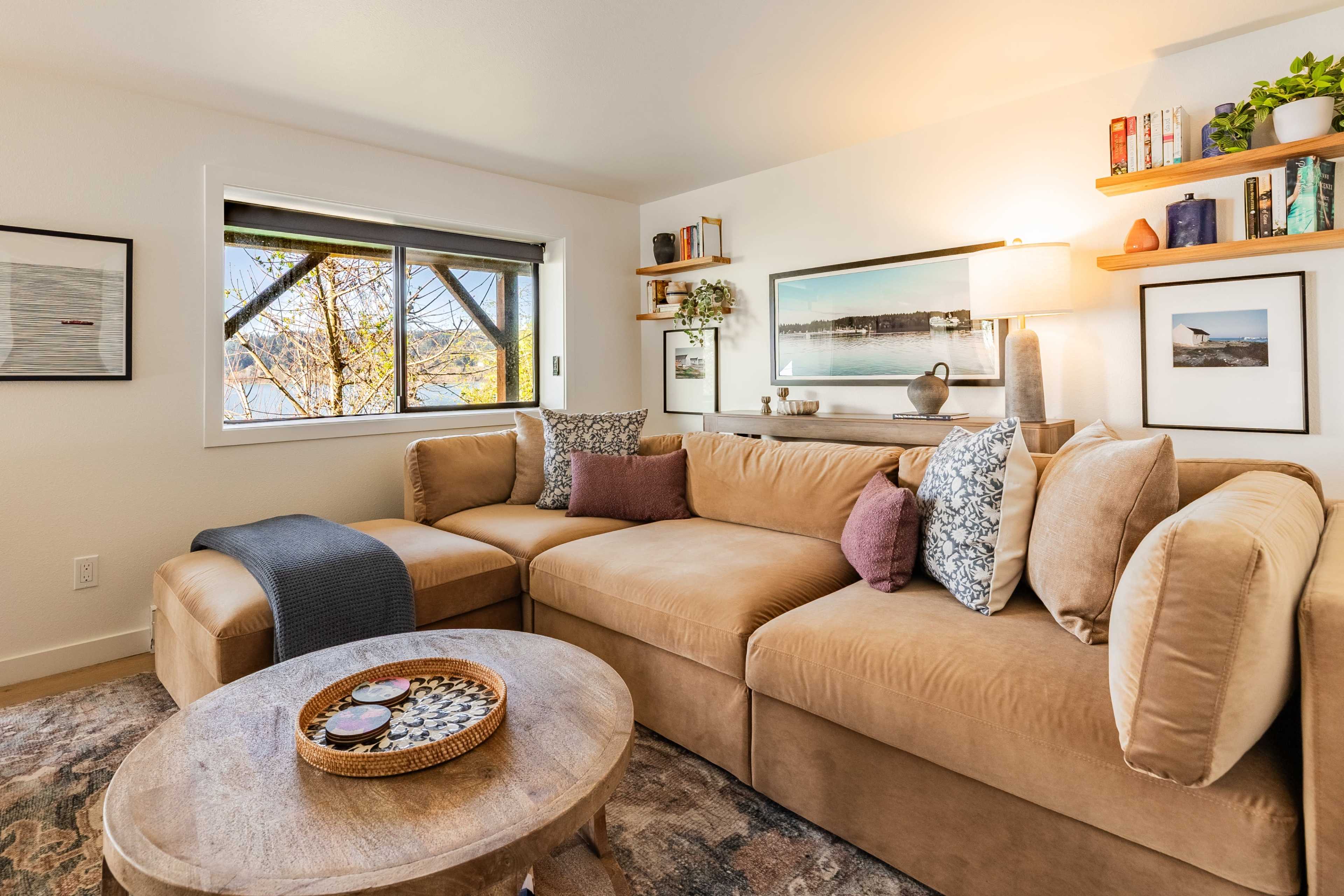 The image shows a cozy living room with a large, light brown sectional sofa, a round coffee table, and a window overlooking a scenic view.