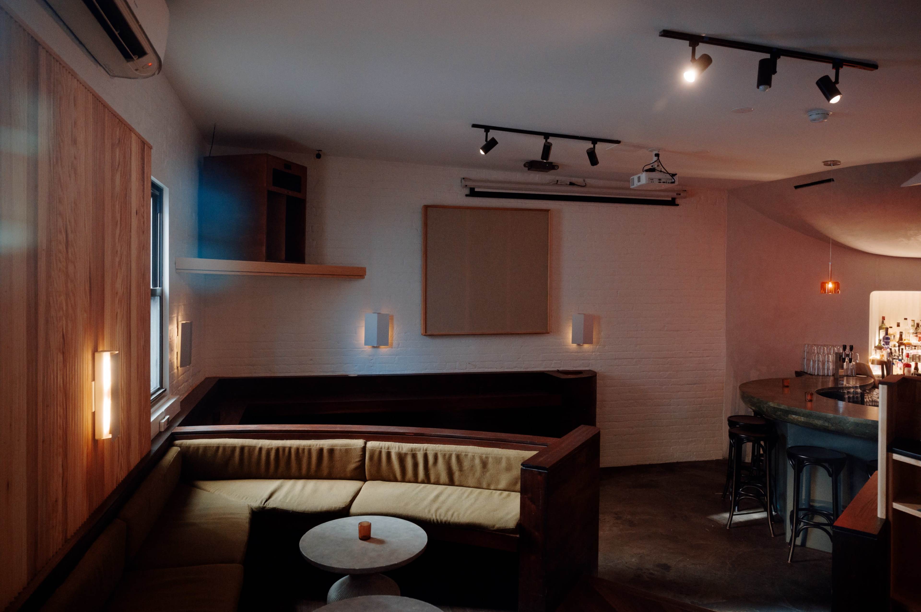 The image shows a dimly lit modern bar interior featuring a curved seating area, a round table, and a bar counter with stools.