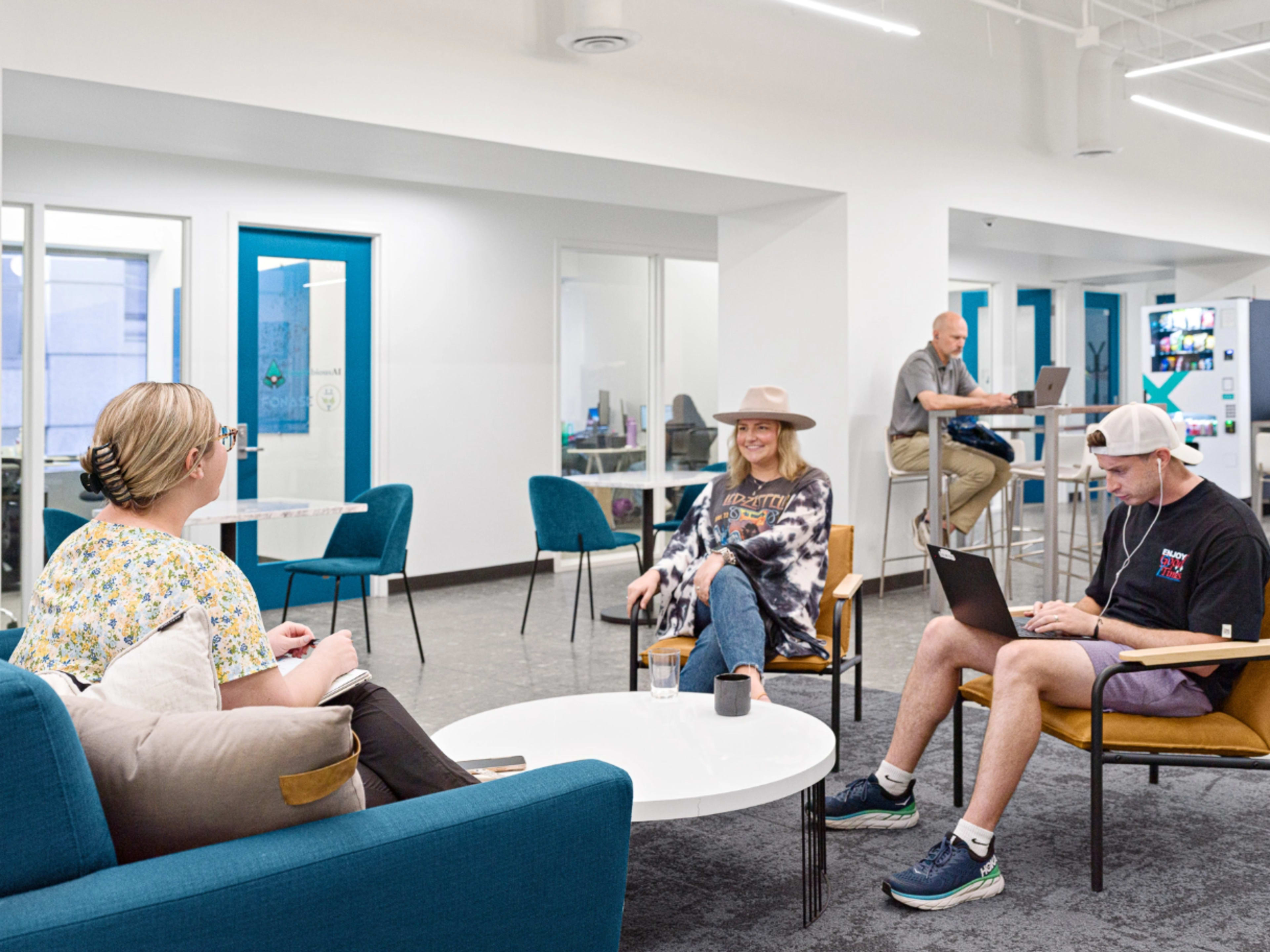 Three people interact in a modern office space, with one sitting at a table using a laptop, another seated in a chair wearing a hat, and a third person standing near a kitchen area.