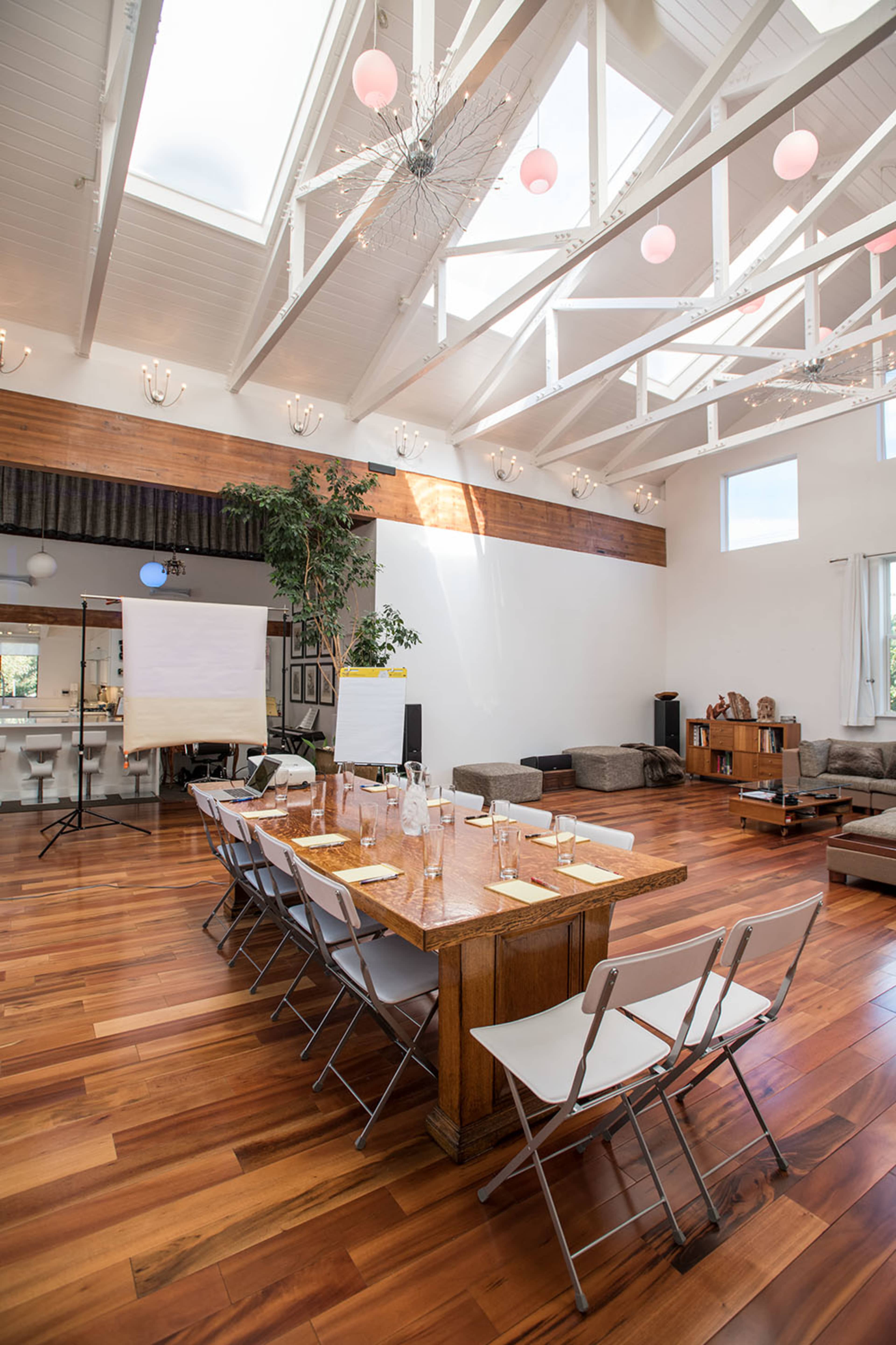 A spacious meeting room features a long wooden table set with plates and glasses, surrounded by chairs, with large windows and a vaulted ceiling adorned with modern light fixtures.