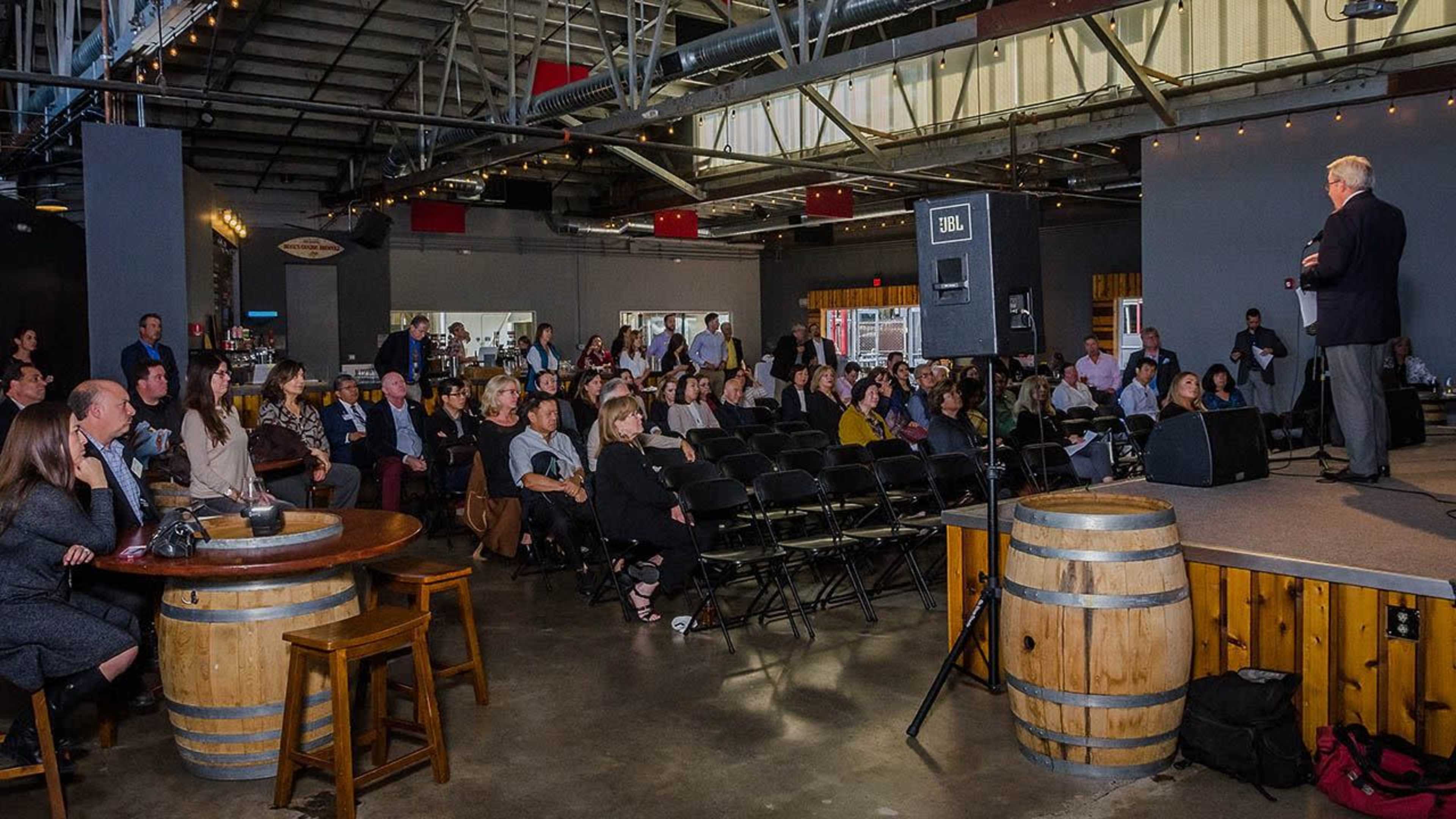 A speaker addresses an audience seated in a spacious venue with wooden barrels and a bar in the foreground.