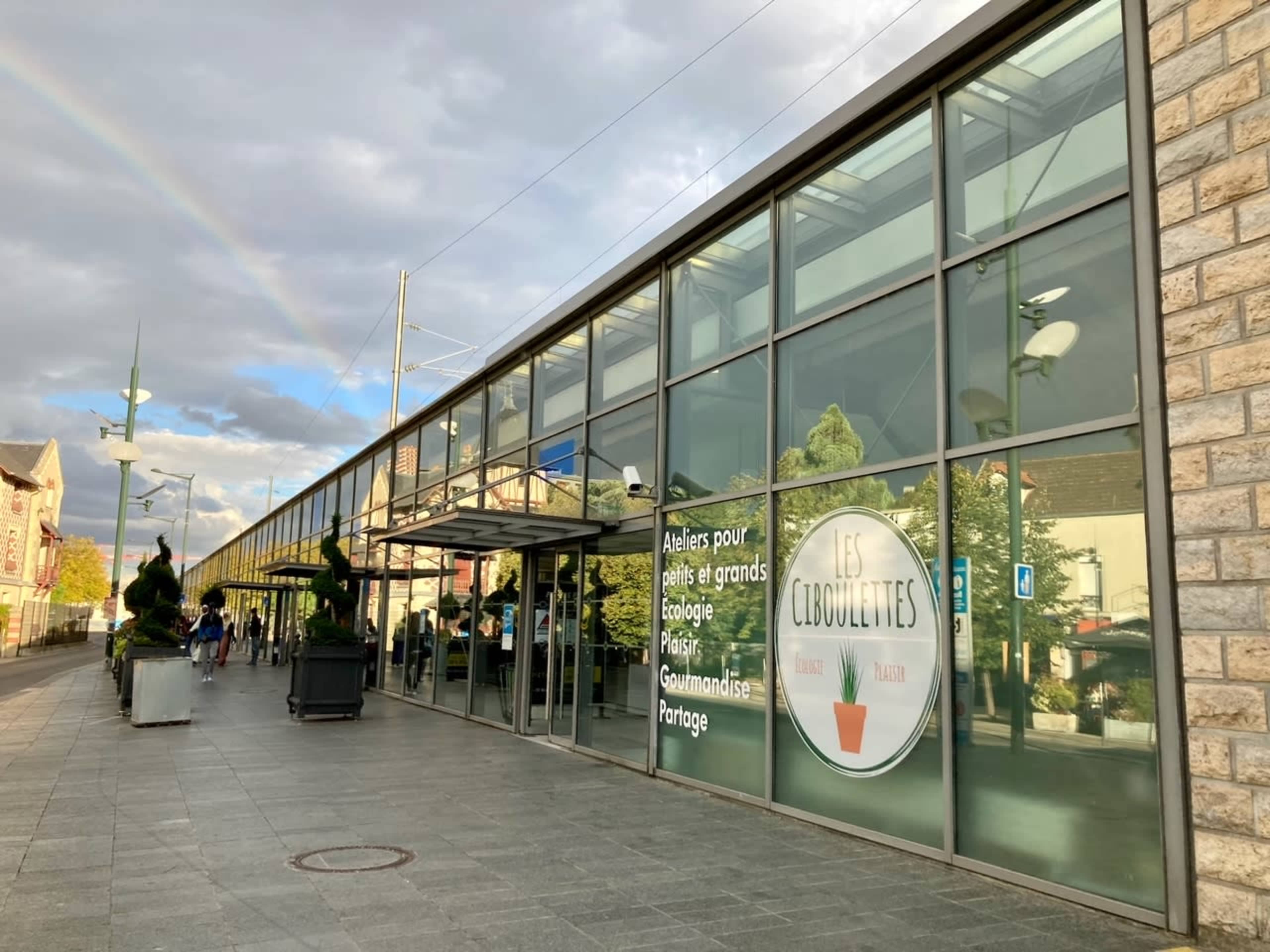A large, glass-fronted building with a sign reading "Les Libouclettes," surrounded by planters and a rainbow in the background.