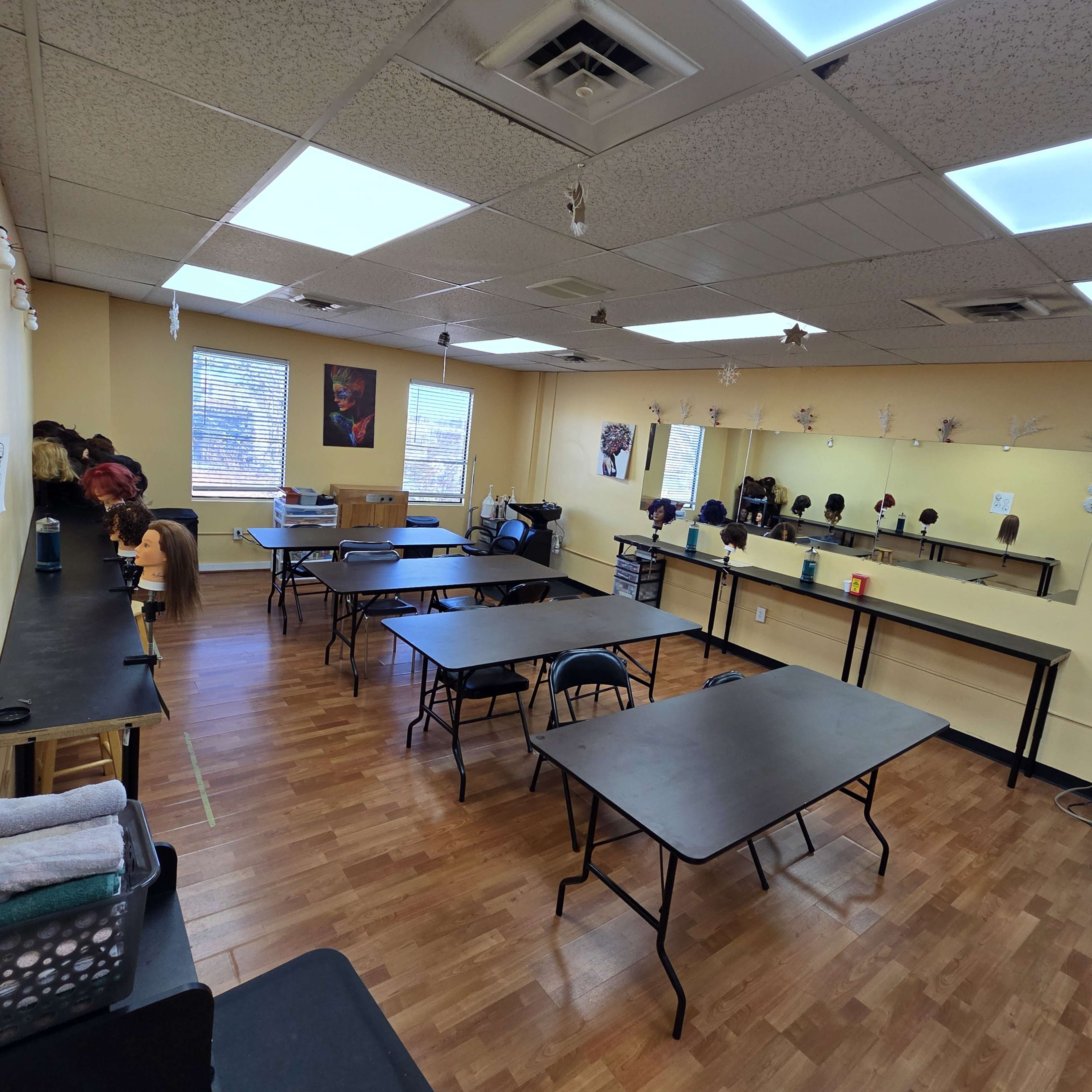 The image shows a spacious classroom or workshop with several black folding tables arranged in a grid, chairs around them, and a large mirror along one wall.