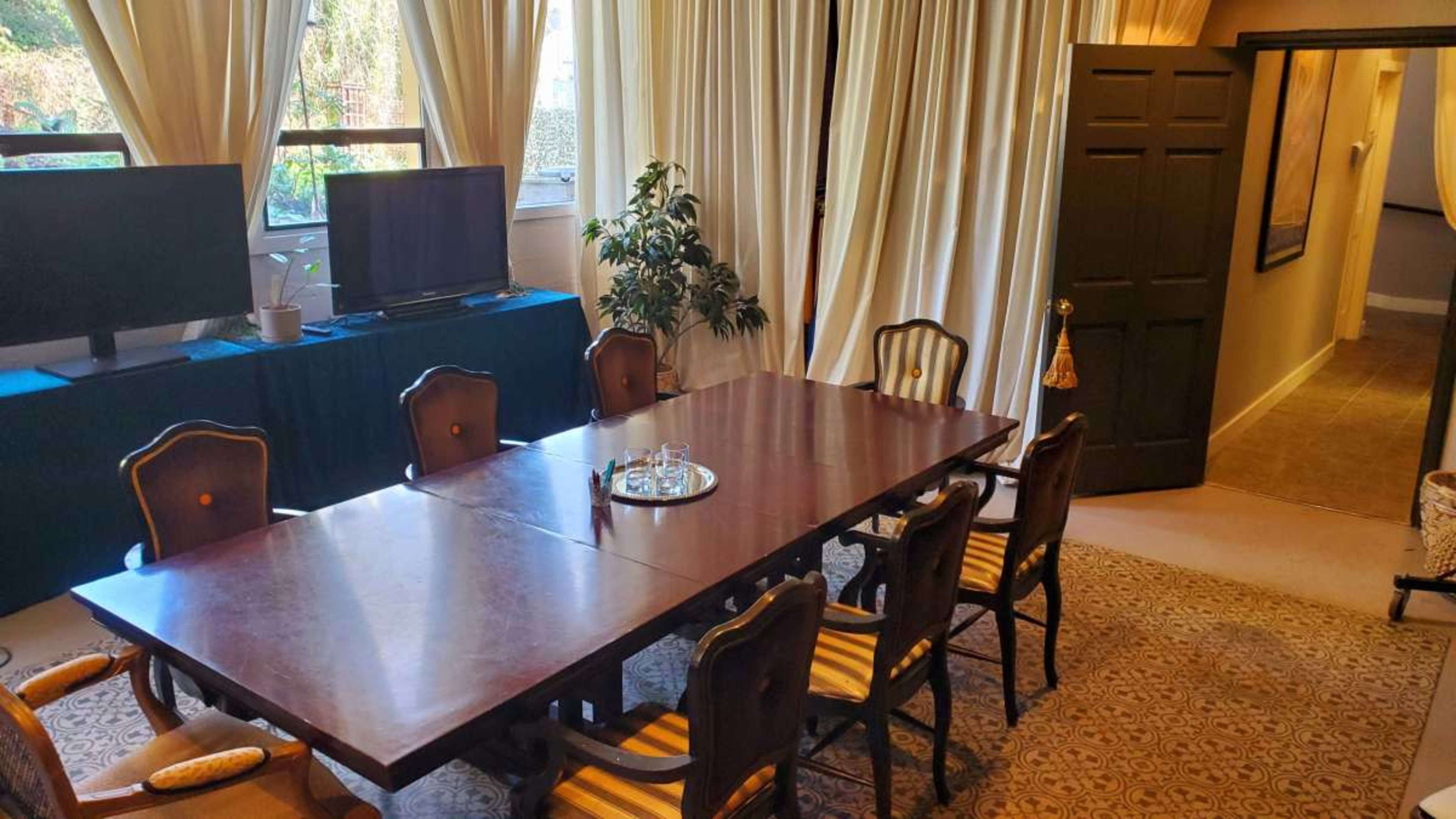 A large wooden dining table accompanied by eight chairs in a room with large windows, plants, and two televisions.