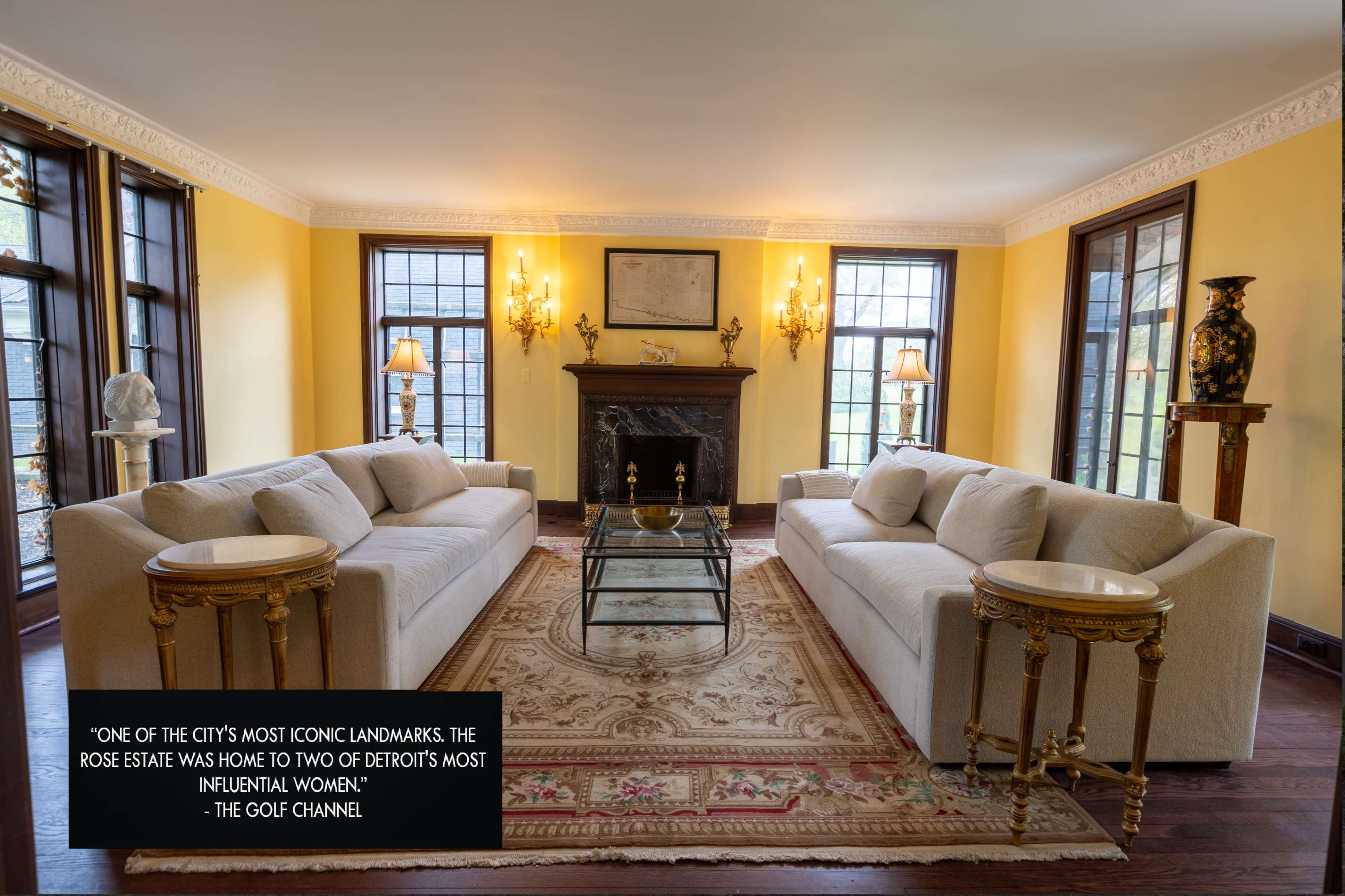 A bright living room with two light-colored sofas facing a glass coffee table in front of a fireplace, featuring ornate decor and large windows.