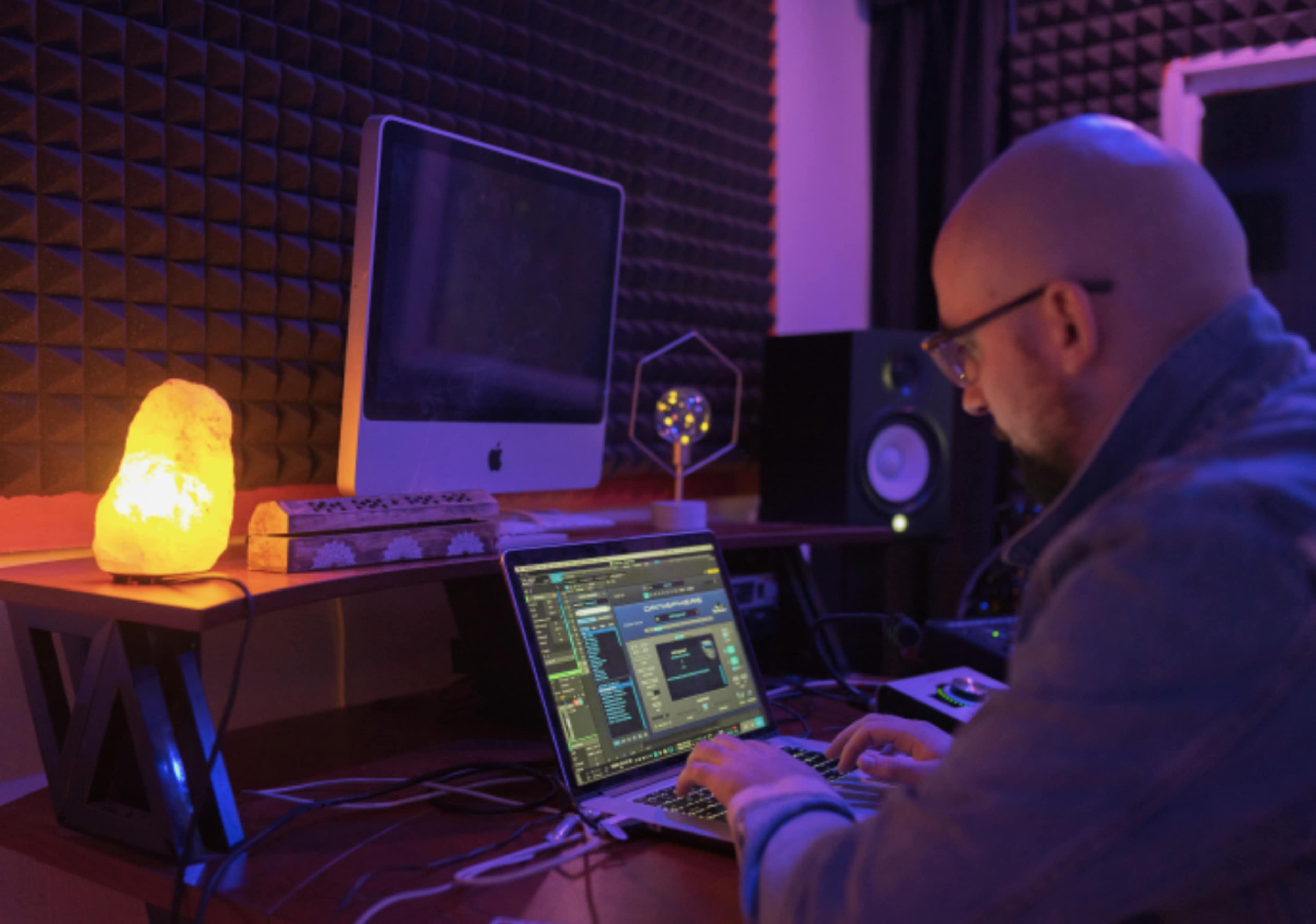 A person works on a laptop in a music studio with acoustic panels and a glowing salt lamp nearby.