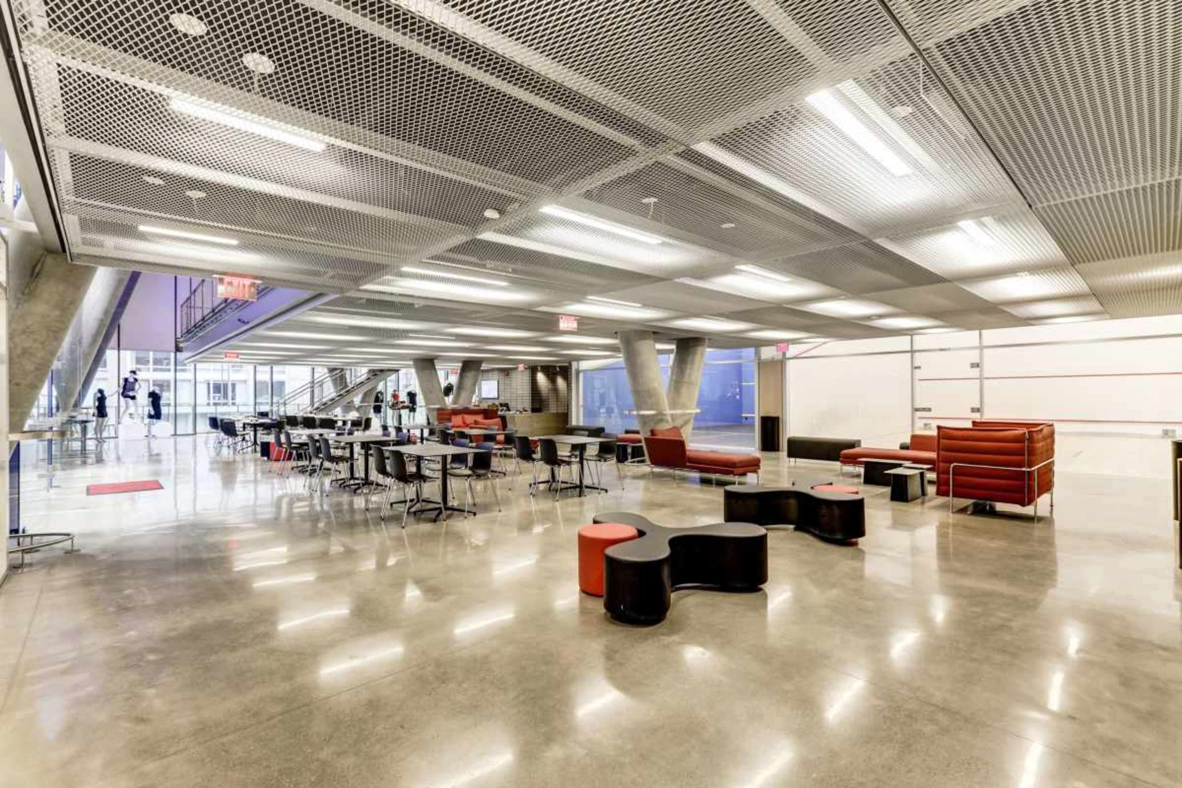 The image shows a spacious, modern interior with seating areas, tables, and a polished concrete floor, featuring geometric lighting and structural elements.