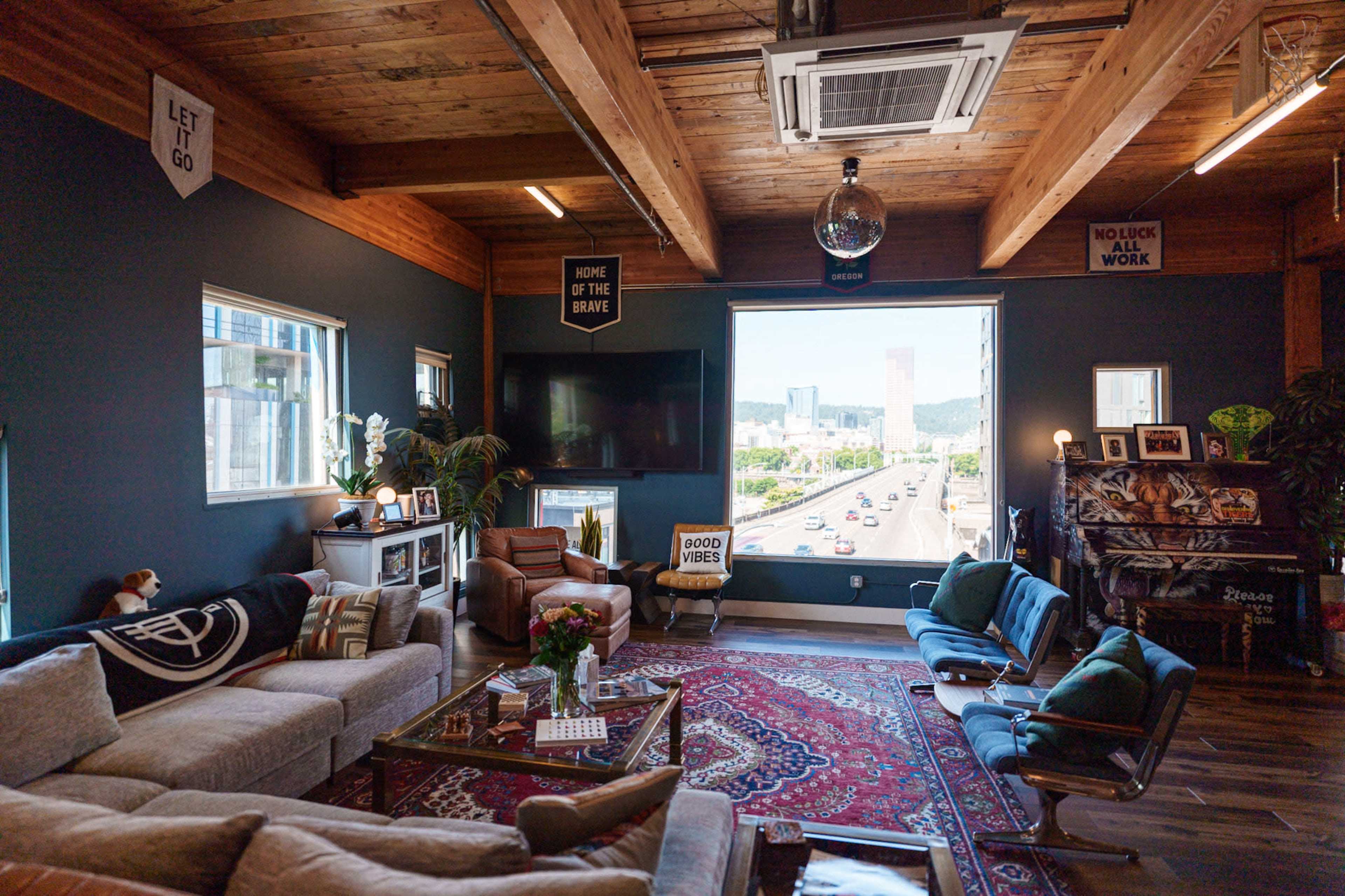 The image shows a modern living room with wooden beams, a large window overlooking a cityscape, and a mix of comfortable seating arrangements around a decorative rug.
