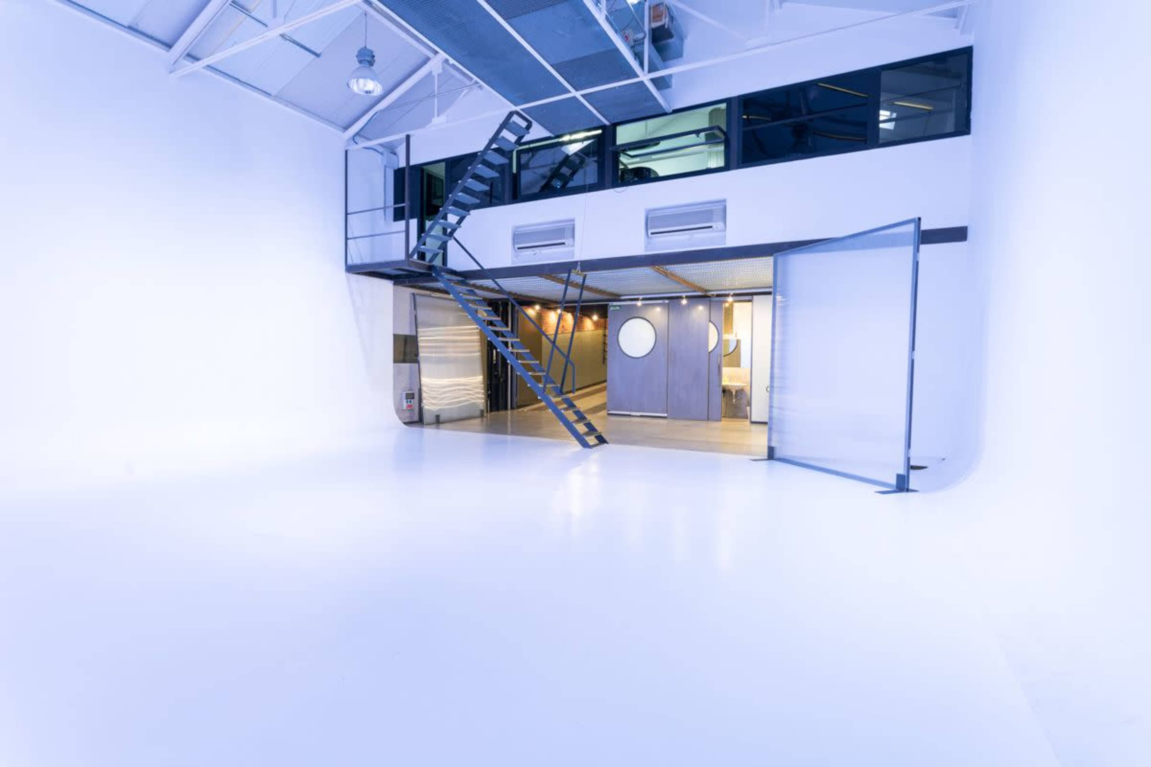 The image shows a spacious, minimalistic studio space with a staircase leading to a second level and large windows allowing natural light.