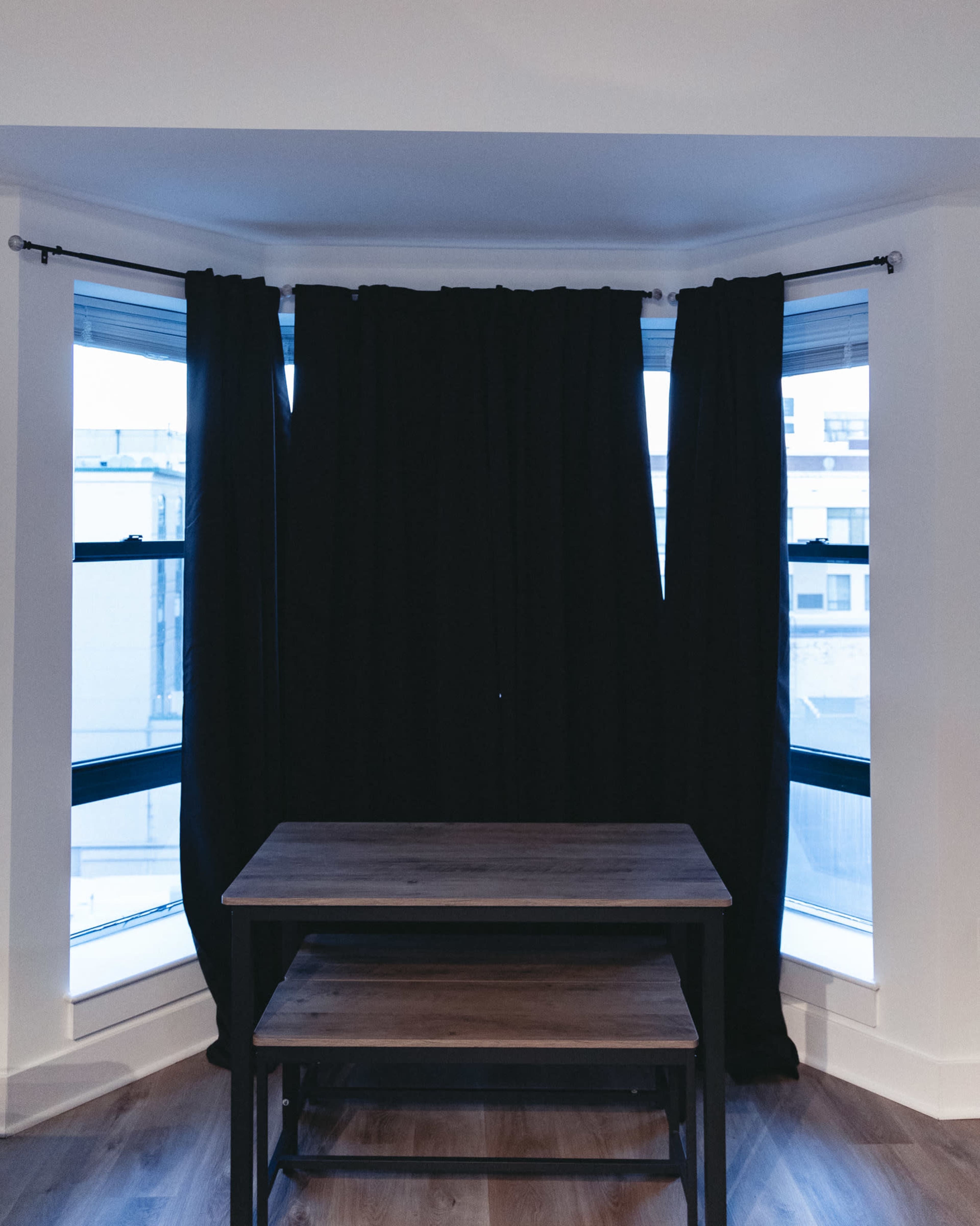 A wooden table is positioned beneath large black curtains in a bright room with windows.