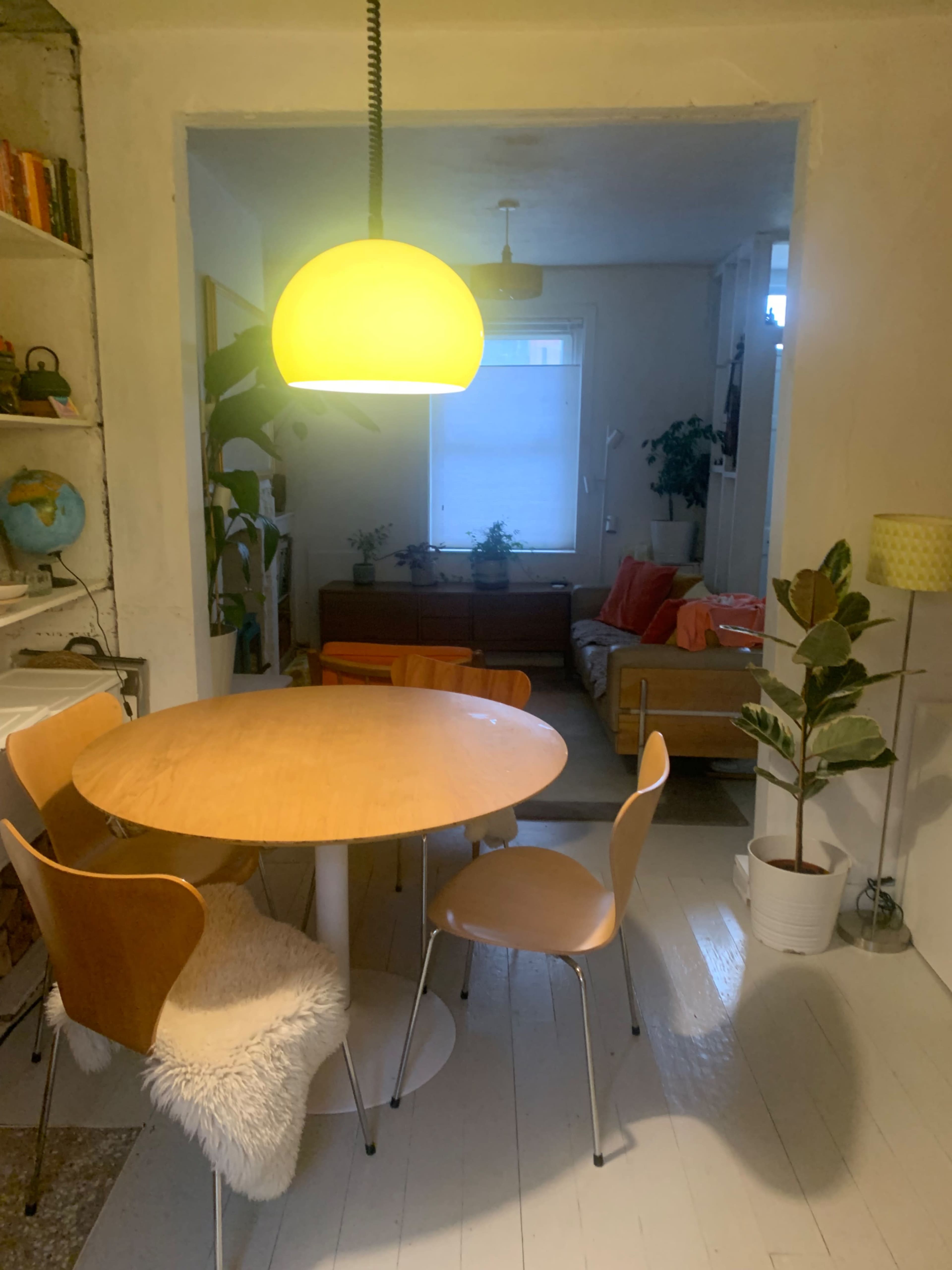 A circular wooden dining table with three chairs is positioned in a light-filled room that connects to a living area furnished with a couch and plants.