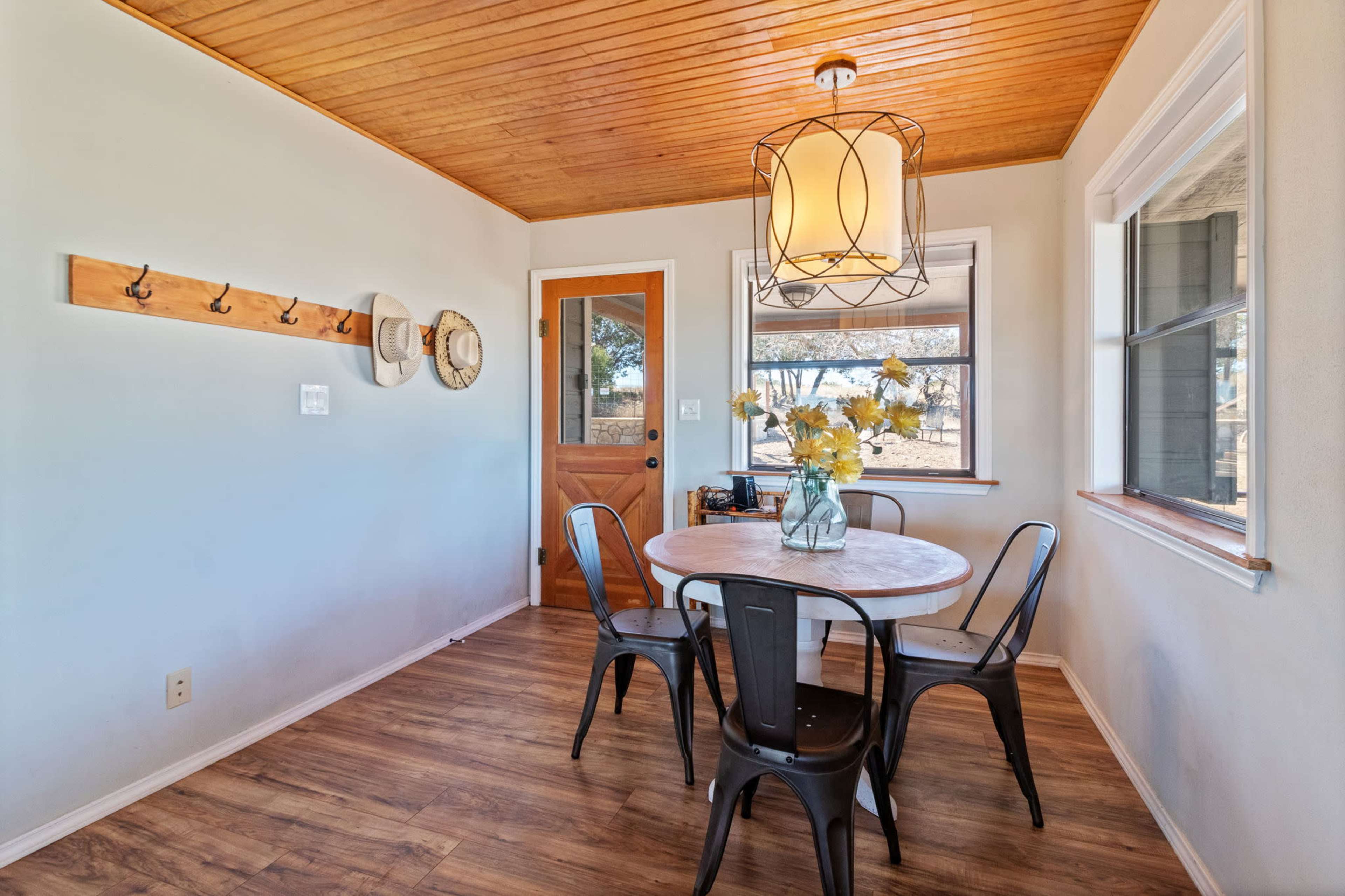 A small dining area features a round table surrounded by black chairs, with a wooden ceiling and a large window letting in natural light.