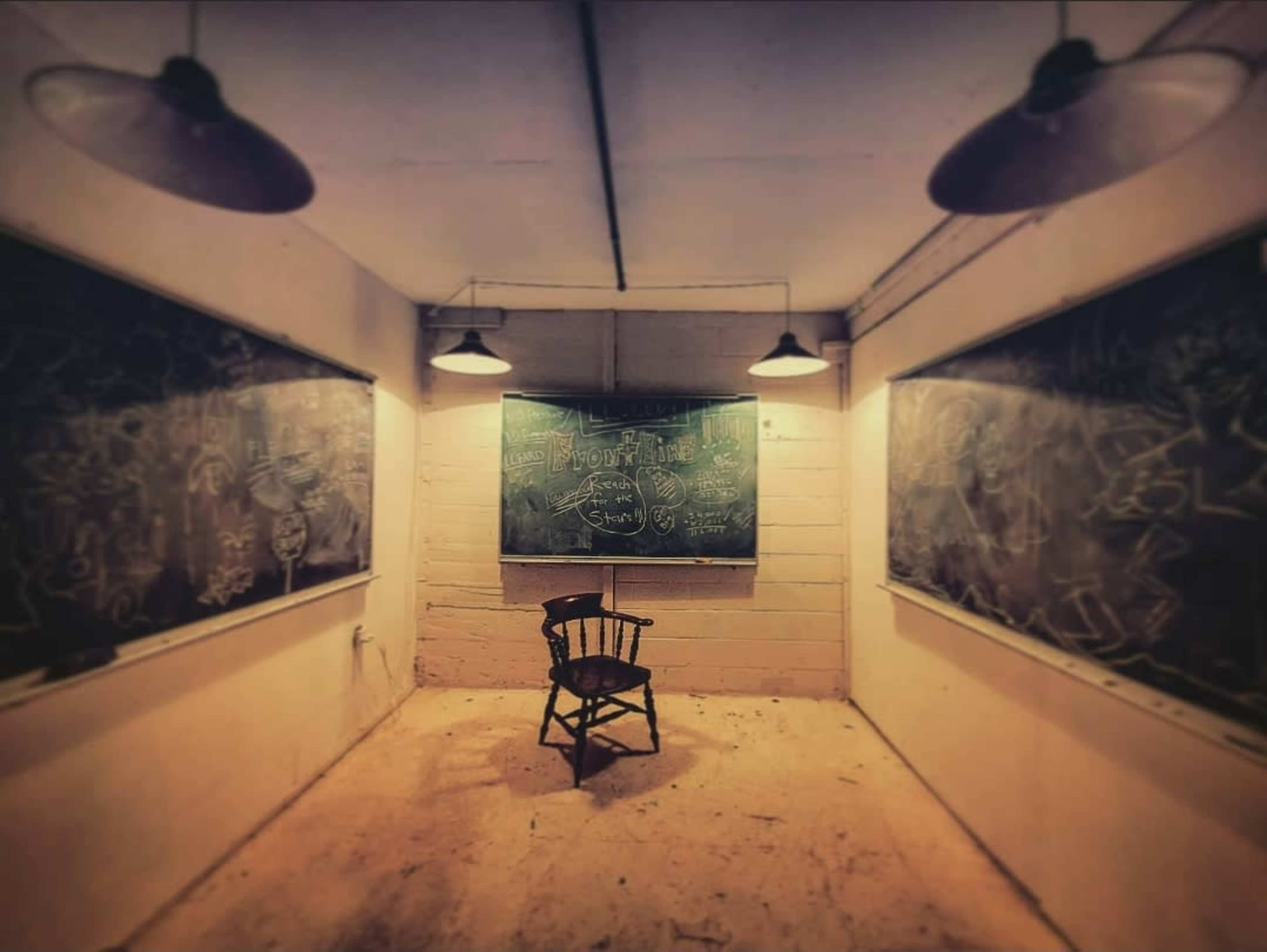 An empty, dimly lit classroom with two chalkboards on either side and a solitary wooden chair in the center.