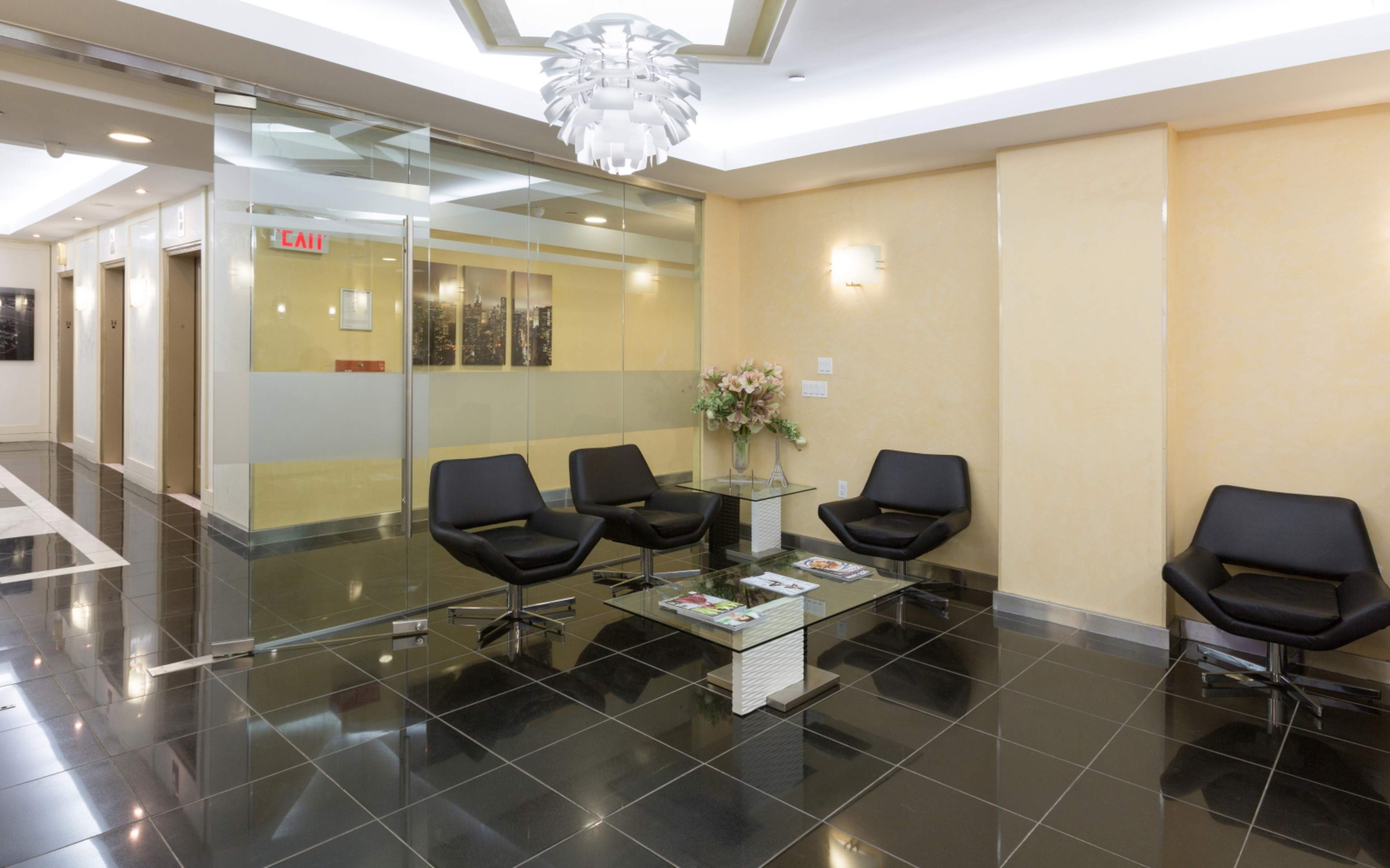 A modern waiting area features black chairs and a glass table, with a flower arrangement and elevator access in the background.
