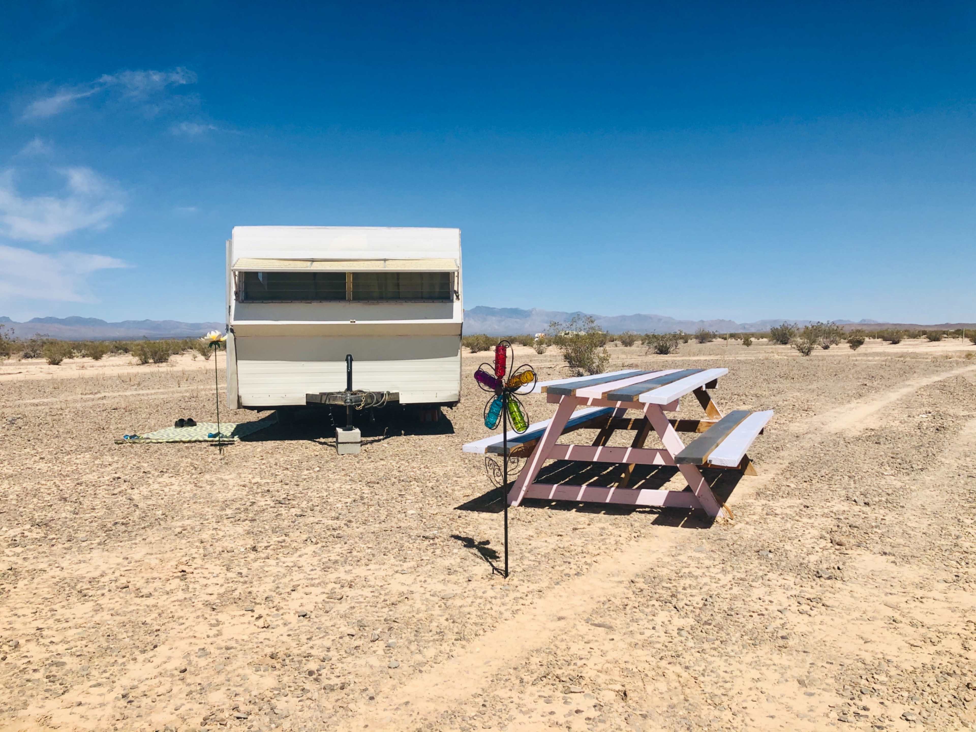 A vintage camper is parked in a barren desert landscape next to a pastel-colored picnic table and a decorative wind spinner.