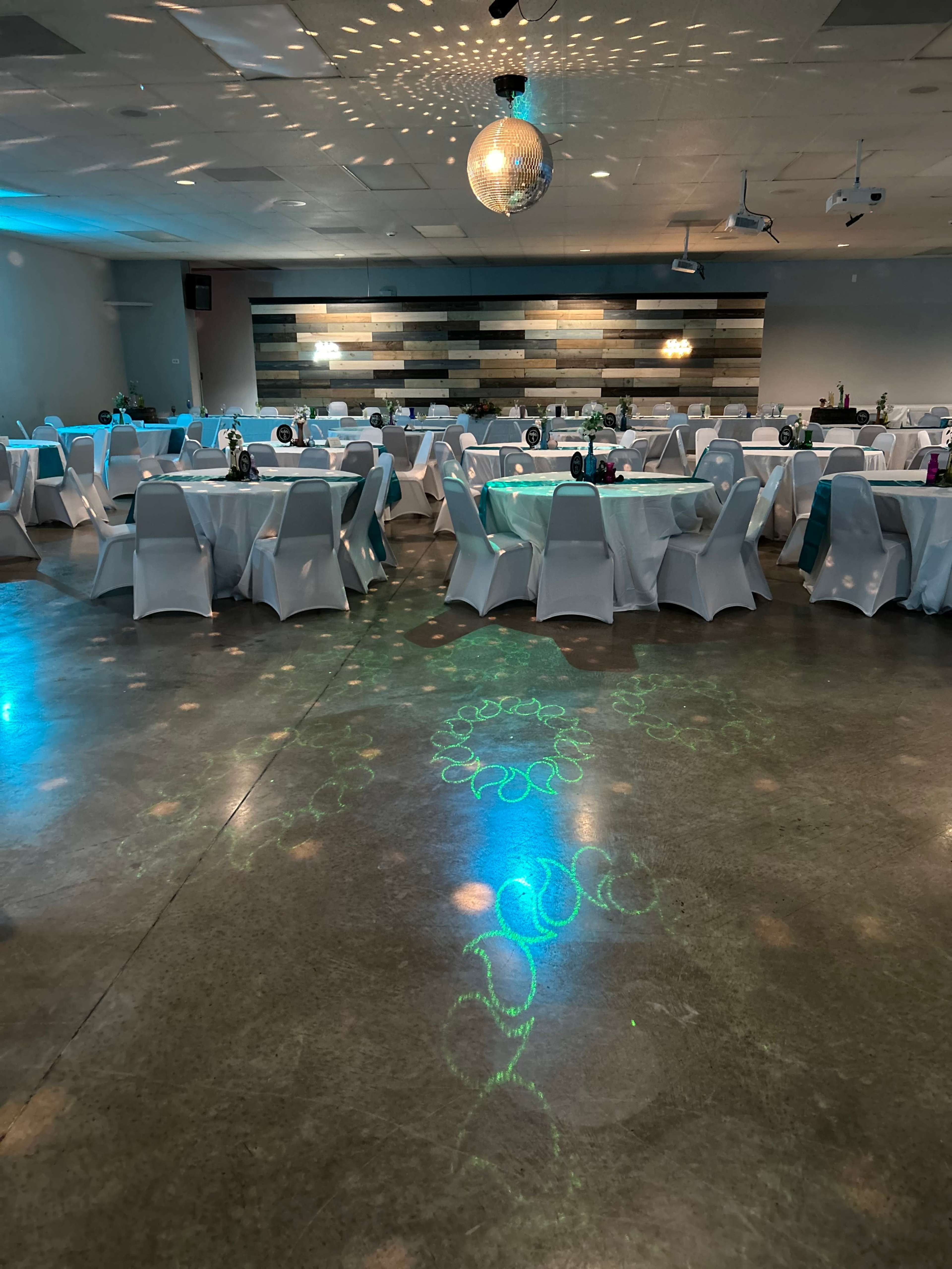 The image shows an event space with round tables covered in white tablecloths, arranged under soft lighting and featuring a disco ball hanging from the ceiling.